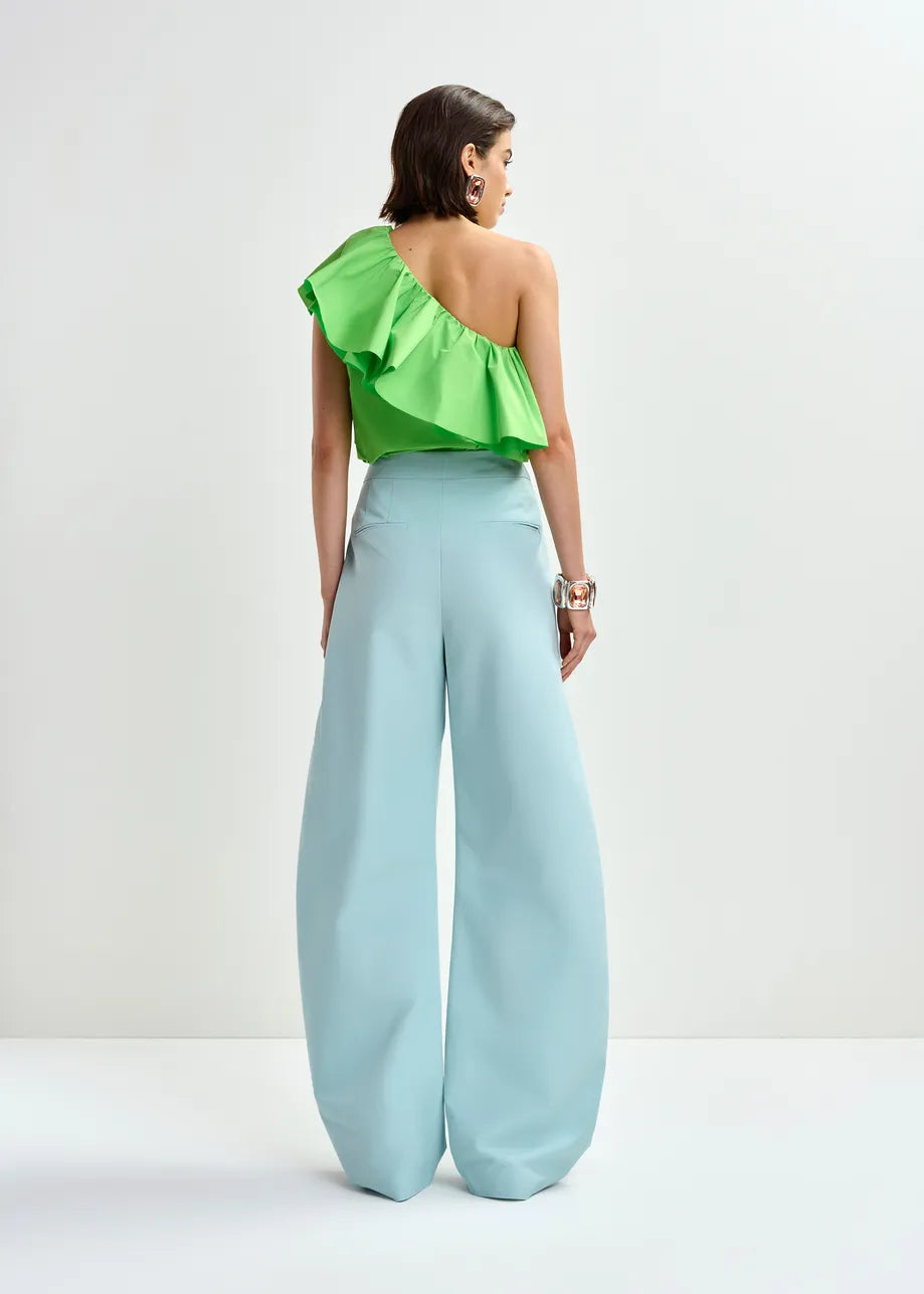 A woman, seen from behind, wears the Essentiel Antwerp Jillian Asymmetric Ruffle Top in green with cascading ruffles and high-waisted wide-leg light blue pants against a plain white background.