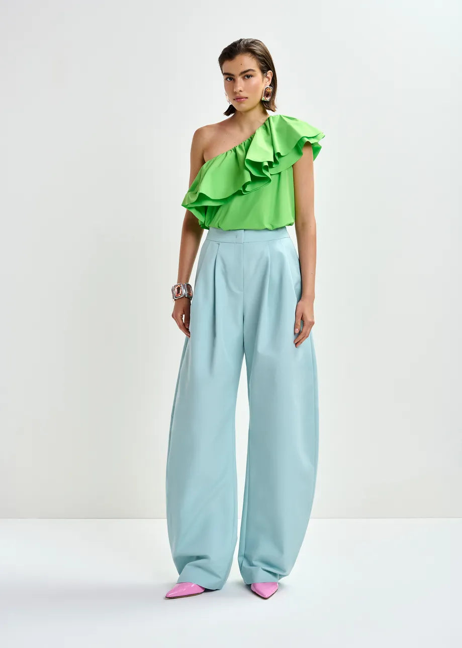 A woman poses against a white background in the Essentiel Antwerp Jillian Asymmetric Ruffle Top in green, paired with light blue wide-leg trousers, pink pointed shoes, and statement jewelry.
