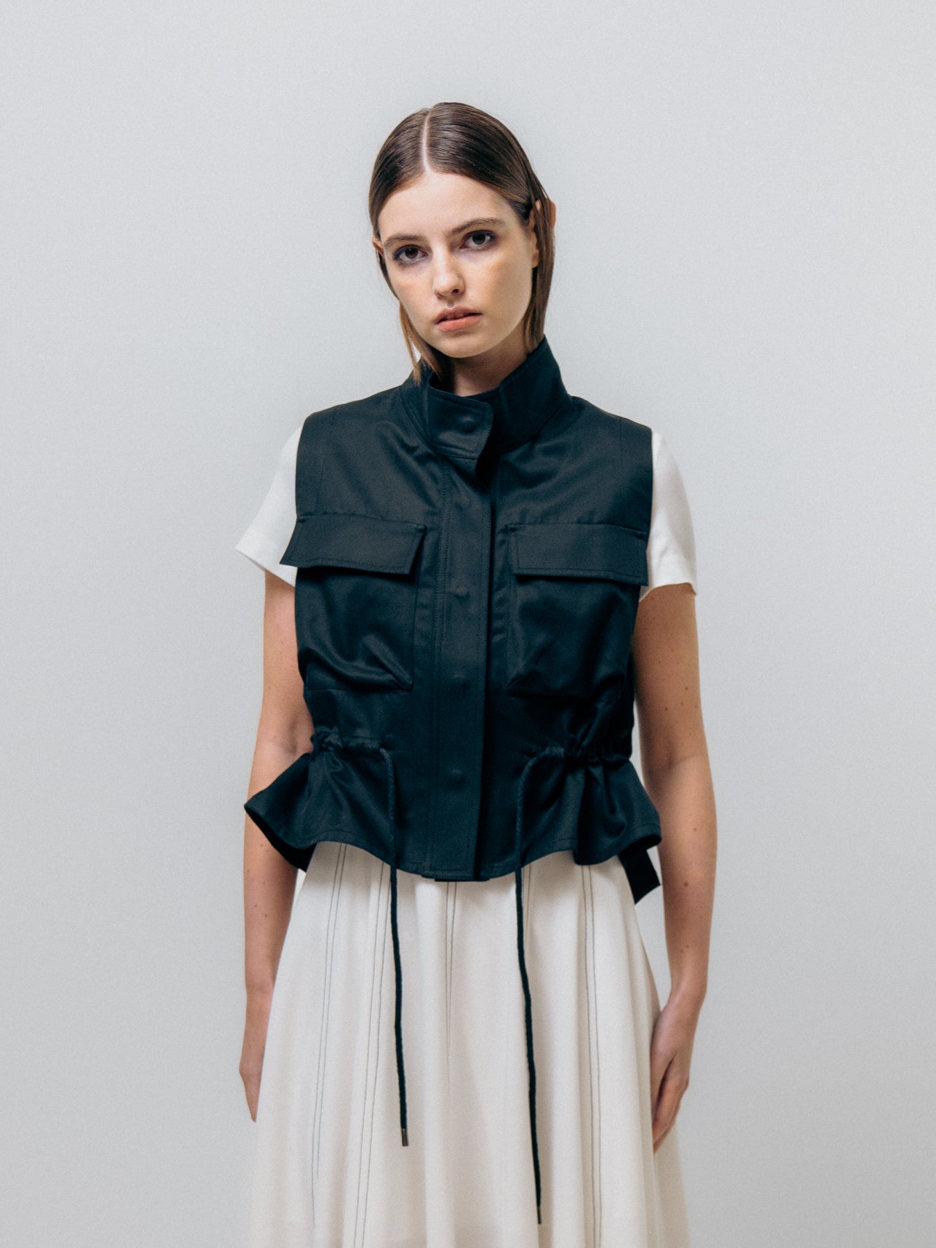 Wearing Emin and Paul's Utility Vest - Black over a white dress, a woman features the chic drawstring waist style as she stands against a plain background, looking at the camera.