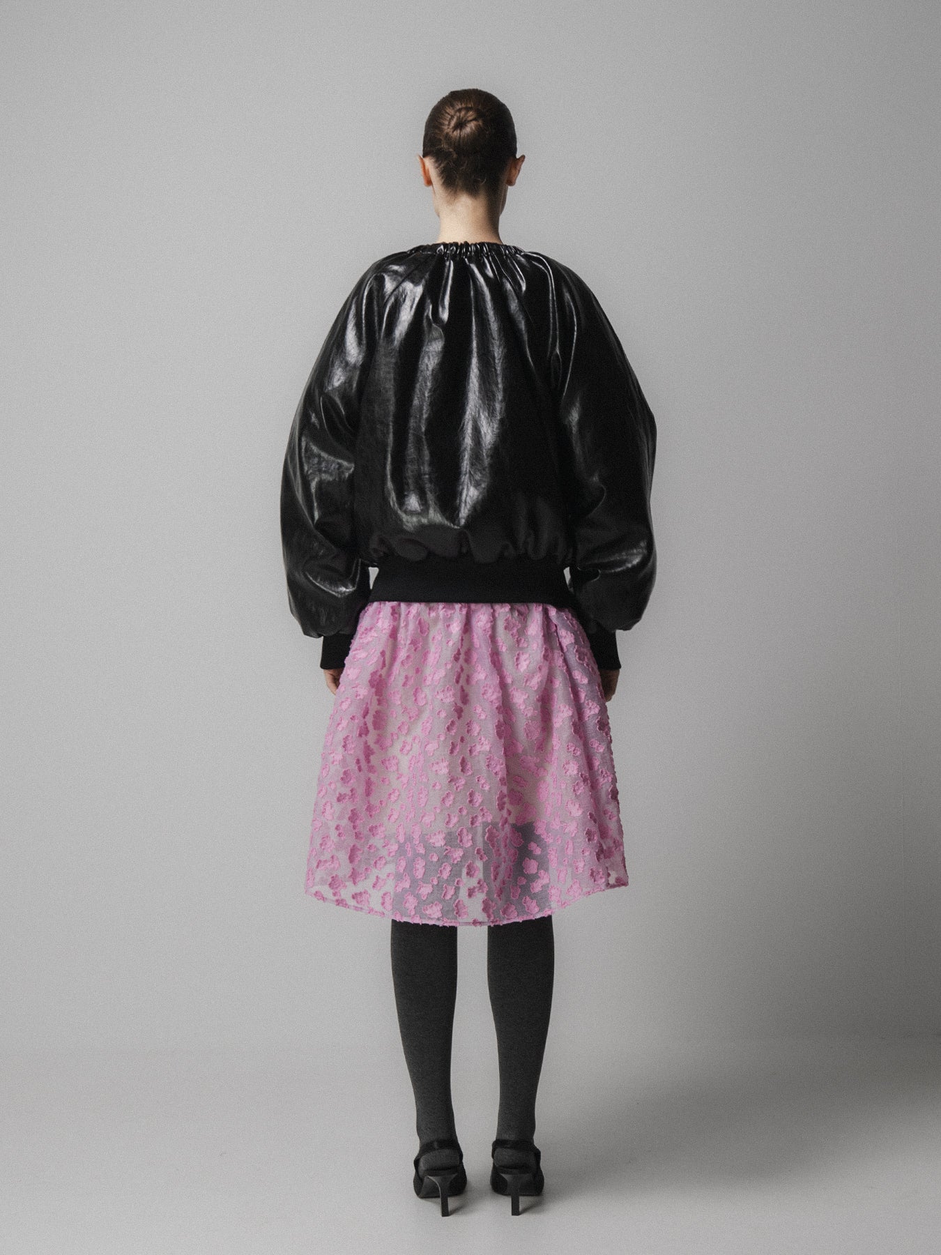 woman wears emin and paul black leather jacket with a pink skirt and grey tights and black shoes on a grey background