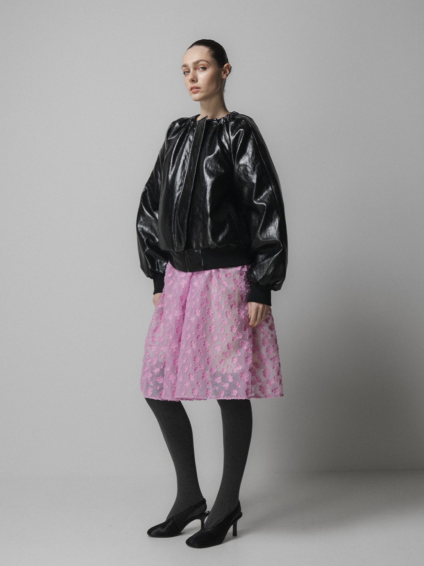woman wears emin and paul black leather jacket with a pink skirt and grey tights and black shoes on a grey background