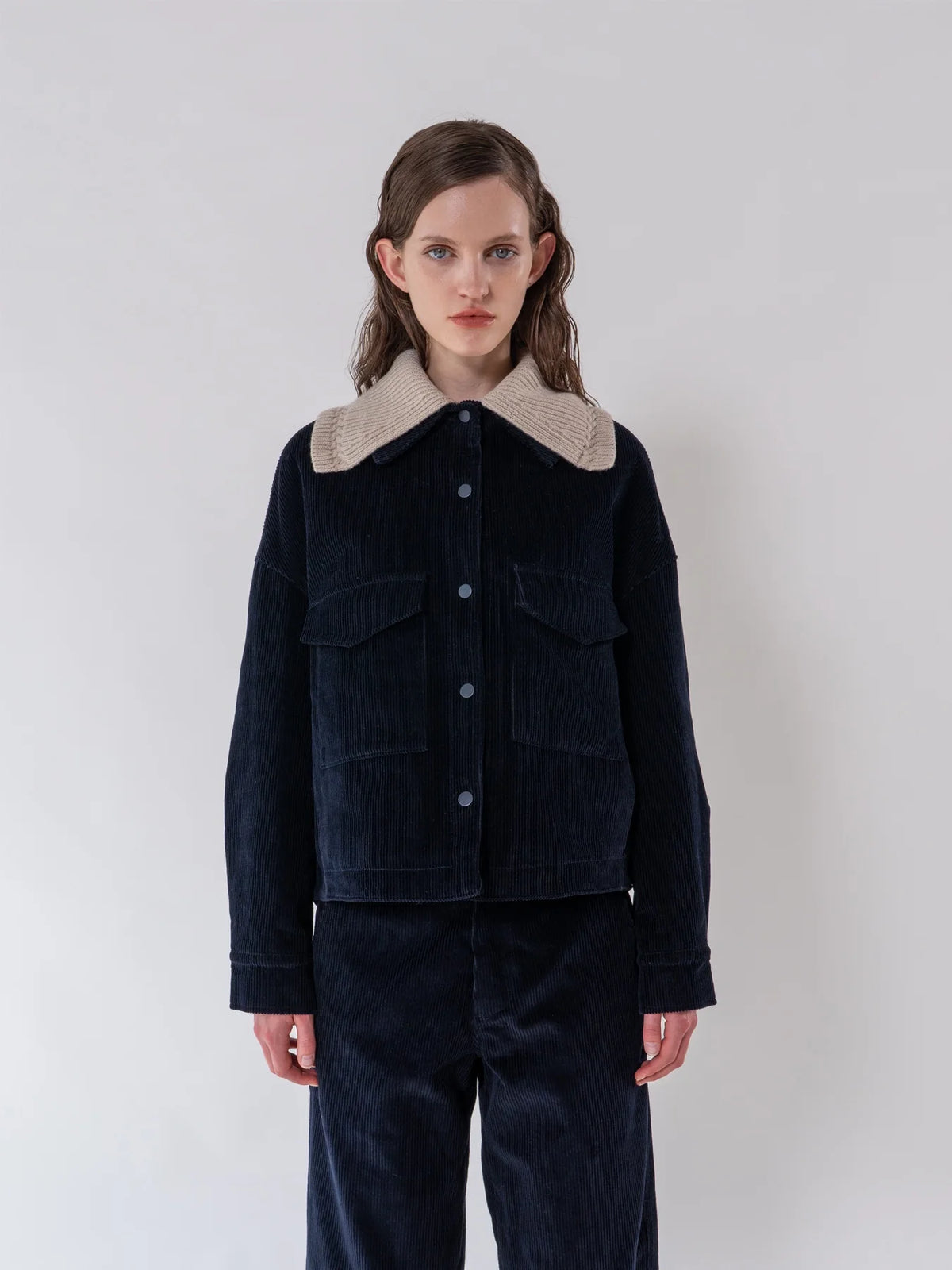 A model wearing a dark navy corduroy jacket with a beige textured collar, paired with matching dark corduroy trousers, against a plain light gray studio background.