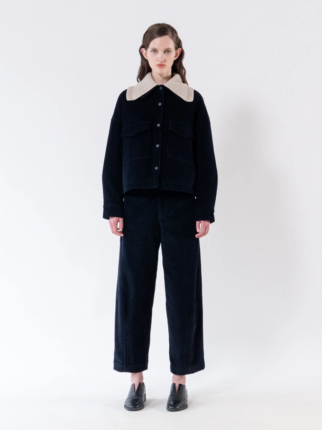 A model stands against a plain white studio background wearing a dark navy corduroy jacket with a contrasting light beige collar, matching dark cropped trousers, and black shoes.