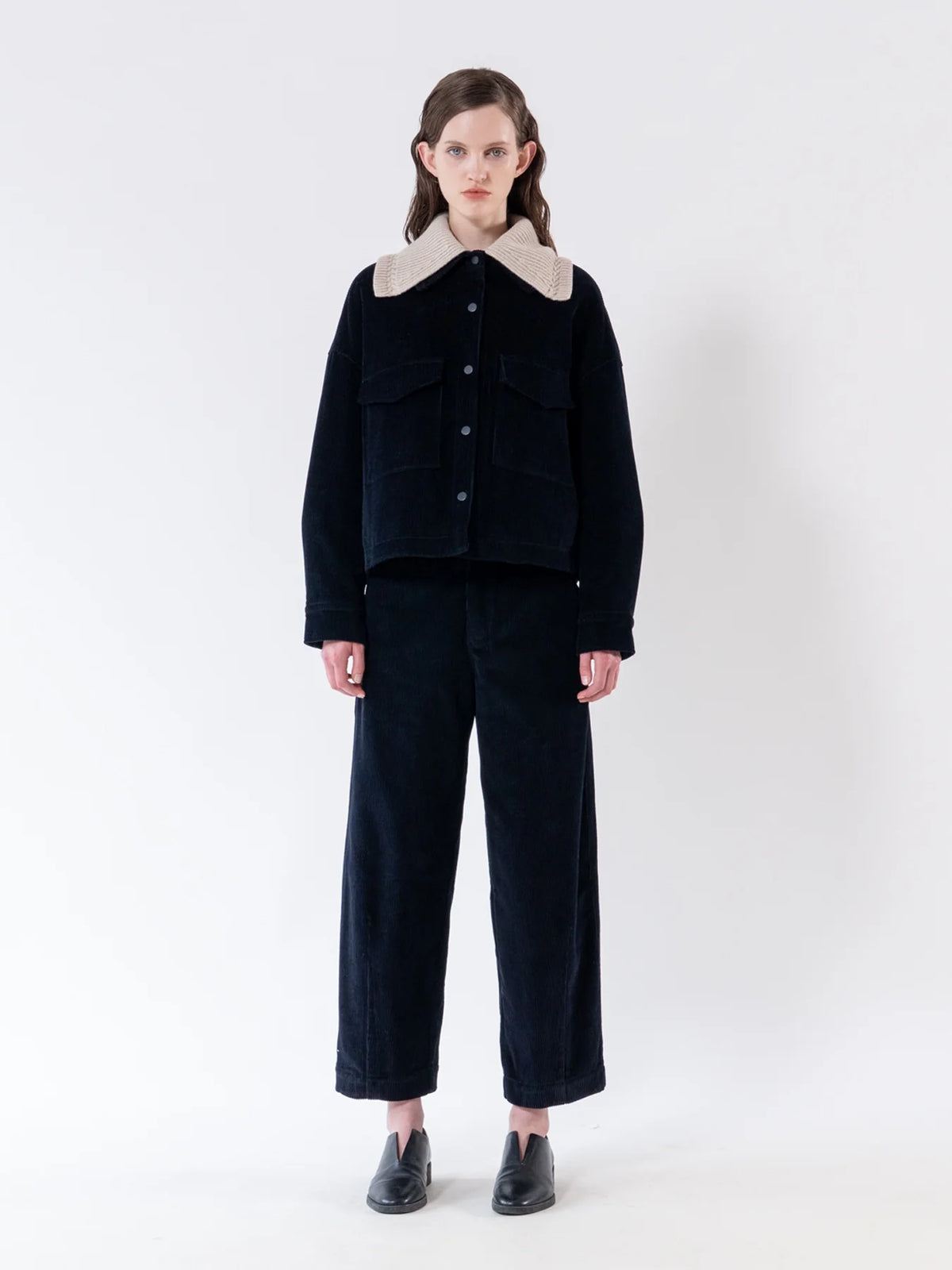 A model stands against a plain white studio background wearing a dark navy corduroy jacket with a contrasting light beige collar, matching dark cropped trousers, and black shoes.