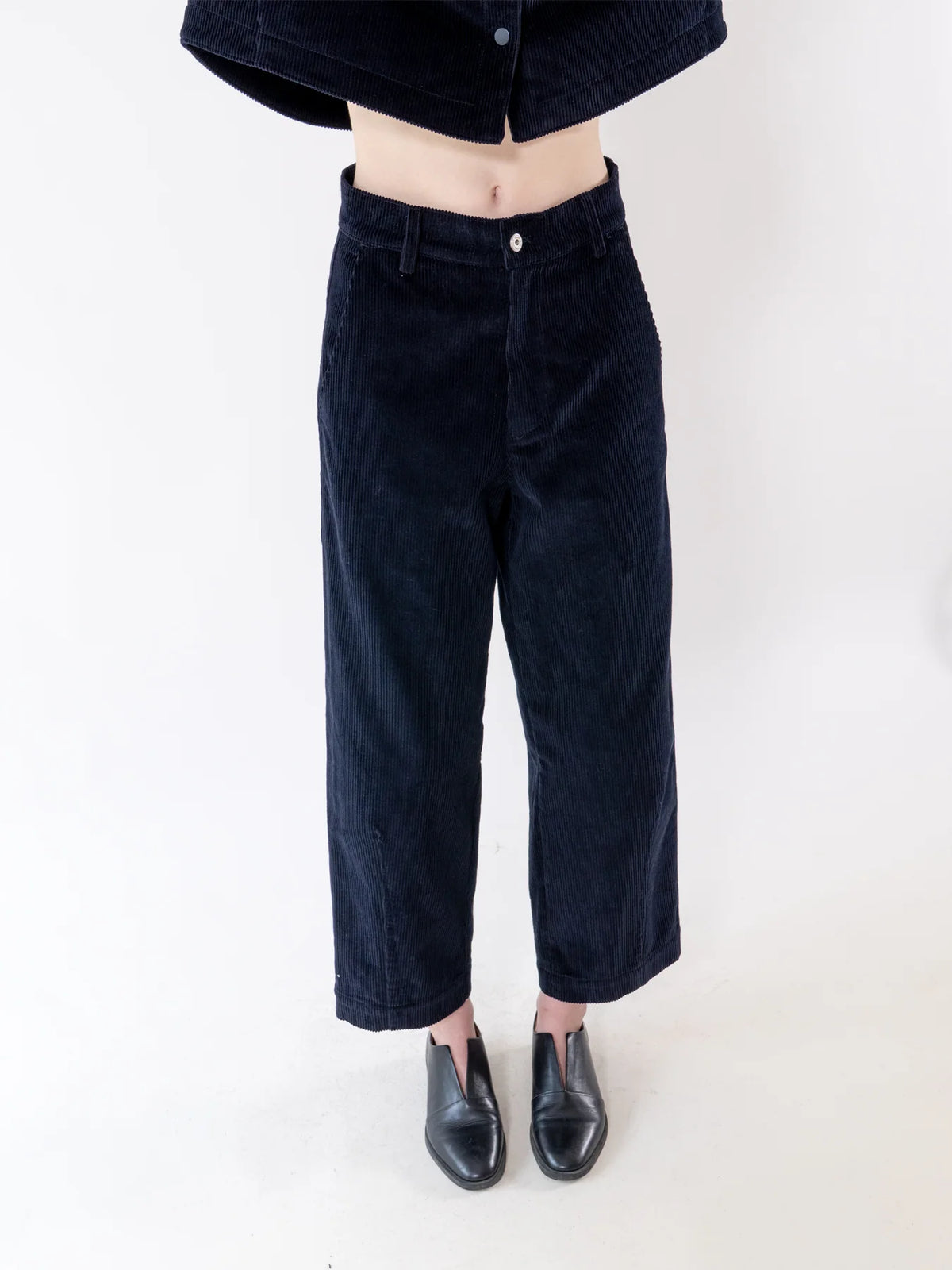 A person wearing dark navy corduroy cropped pants with a cropped matching top and black leather shoes, shown from the waist down against a plain white studio background.