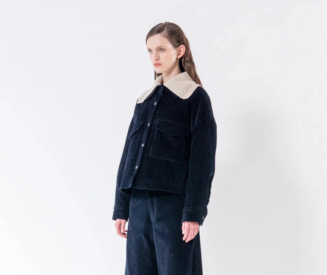 A model stands in a studio wearing a dark navy corduroy jacket with a contrasting beige collar, matching dark navy corduroy trousers, and black leather shoes against a plain white background.
