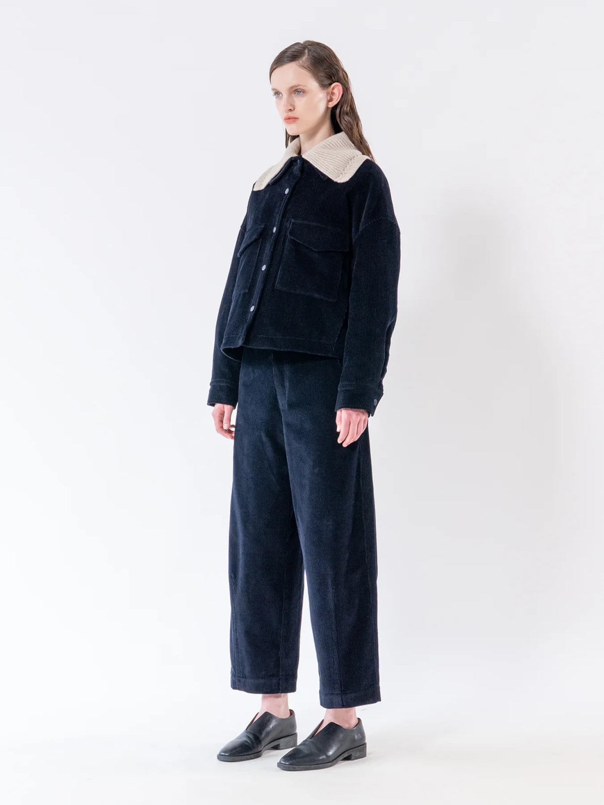 A model stands in a studio wearing a dark navy corduroy jacket with a contrasting beige collar, matching dark navy corduroy trousers, and black leather shoes against a plain white background.