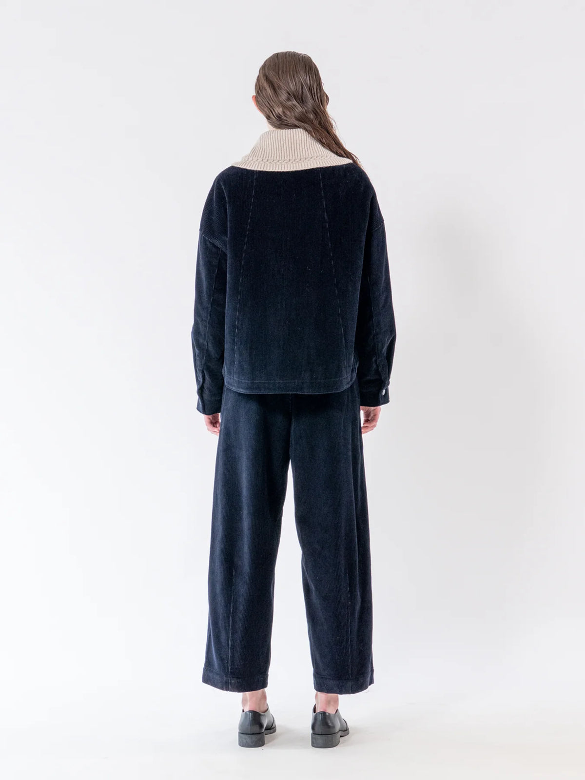 A model viewed from the back wearing a dark navy corduroy two-piece set consisting of a cropped jacket with a cream ribbed knit collar and matching wide-leg trousers, set against a plain white studio background.