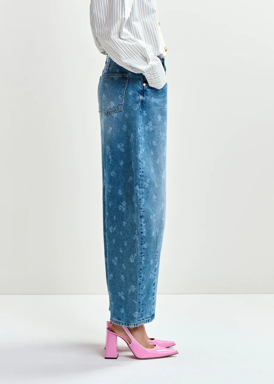 A person stands in profile wearing Essentiel Antwerp’s Jo1 Barrel Cropped Jeans - Blue Floral, paired with a white striped shirt and pink block-heeled shoes against a plain background.