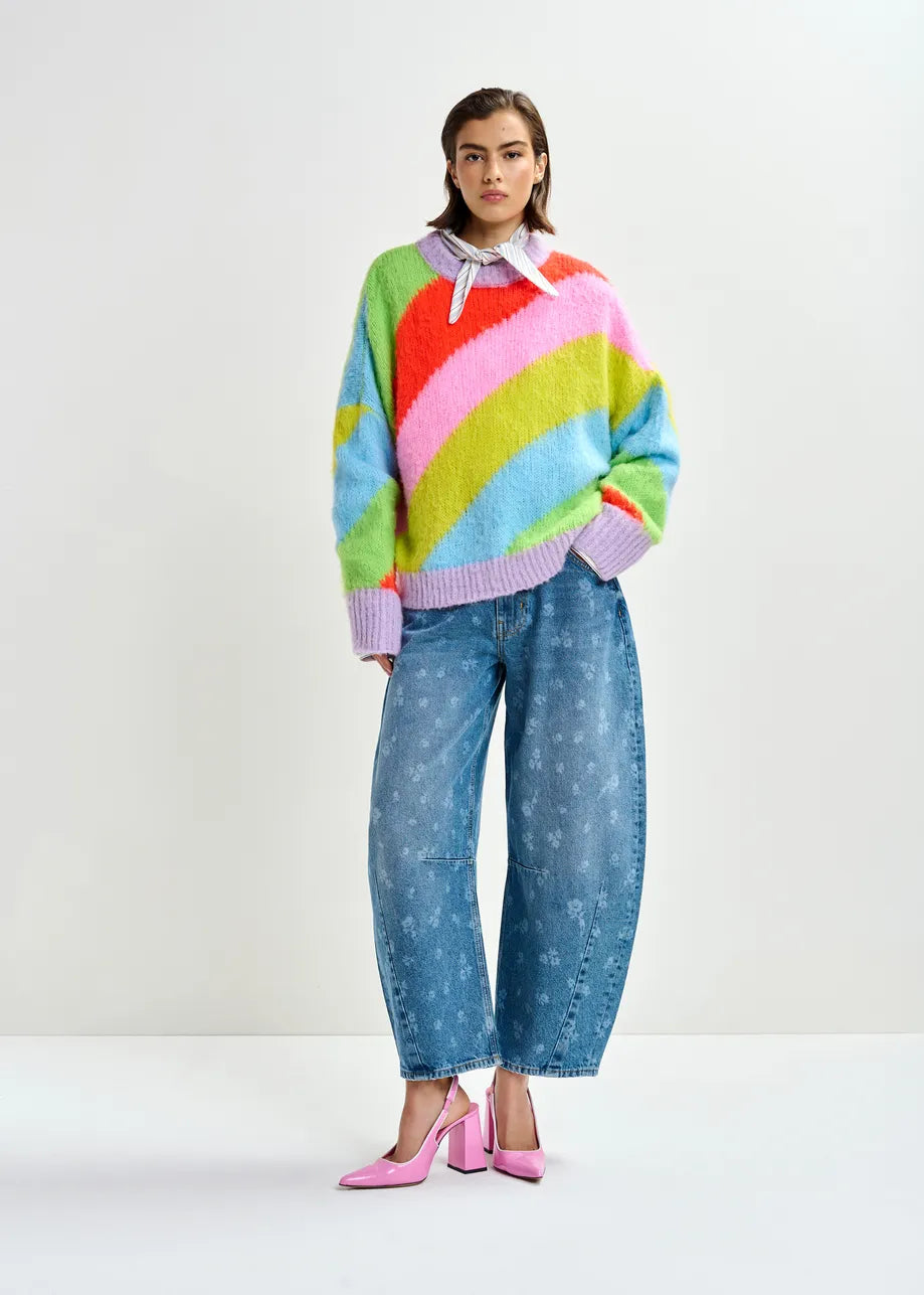 A person stands in a studio wearing Jo1 Barrel Cropped Jeans in blue floral by Essentiel Antwerp, styled with a colorful striped sweater and pink high-heeled shoes.