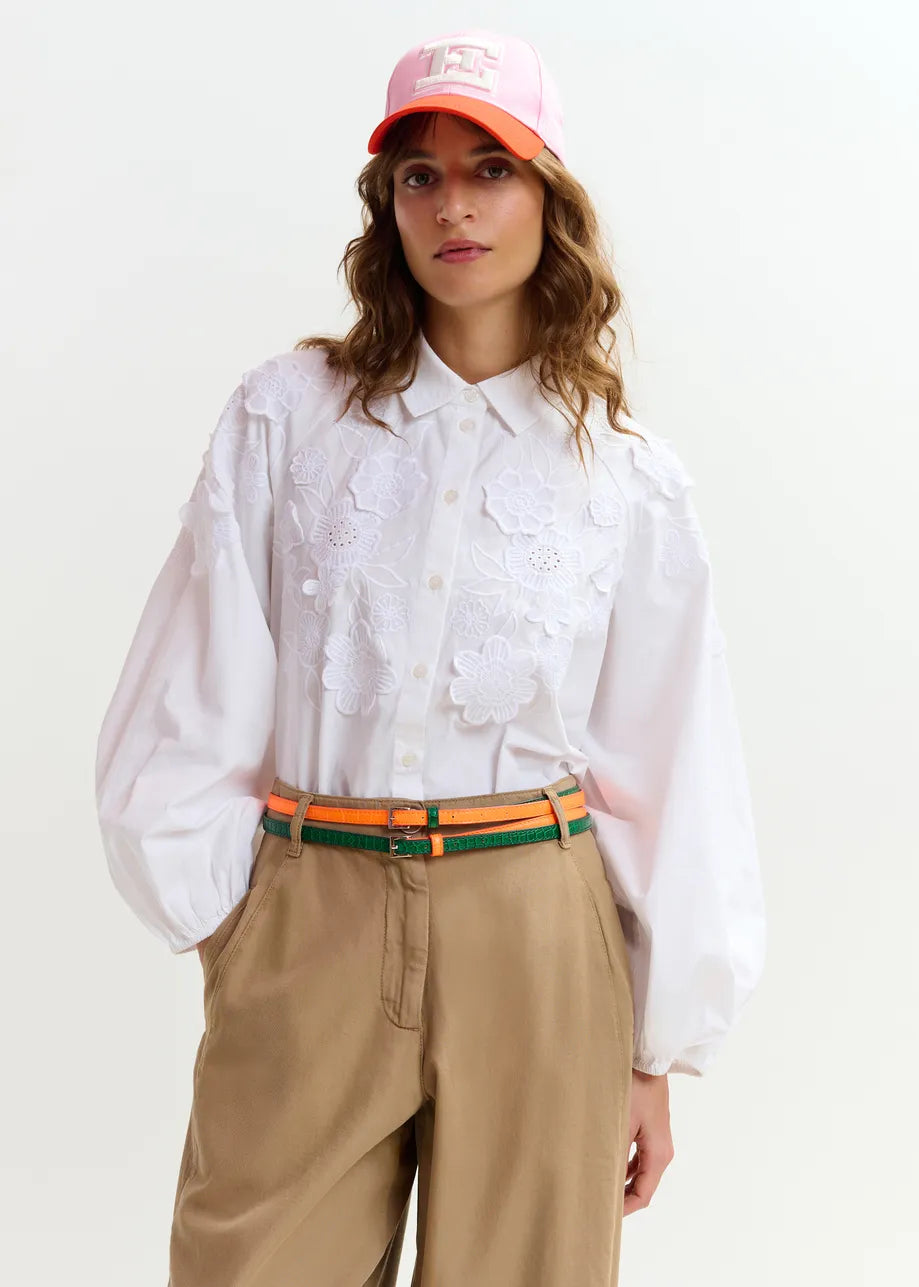 A person wears the Essentiel Antwerp Joella Cotton Flower Shirt in white, featuring floral embroidery and subtle balloon sleeves, along with tan pants with a striped belt and a pink and white baseball cap against a plain background.