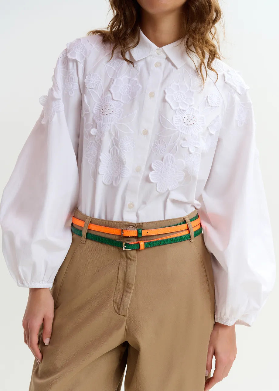 Someone dressed in the Essentiel Antwerp Joella Cotton Flower Shirt in white, featuring long balloon sleeves and floral appliqué, paired with tan pants and an orange-green striped belt.