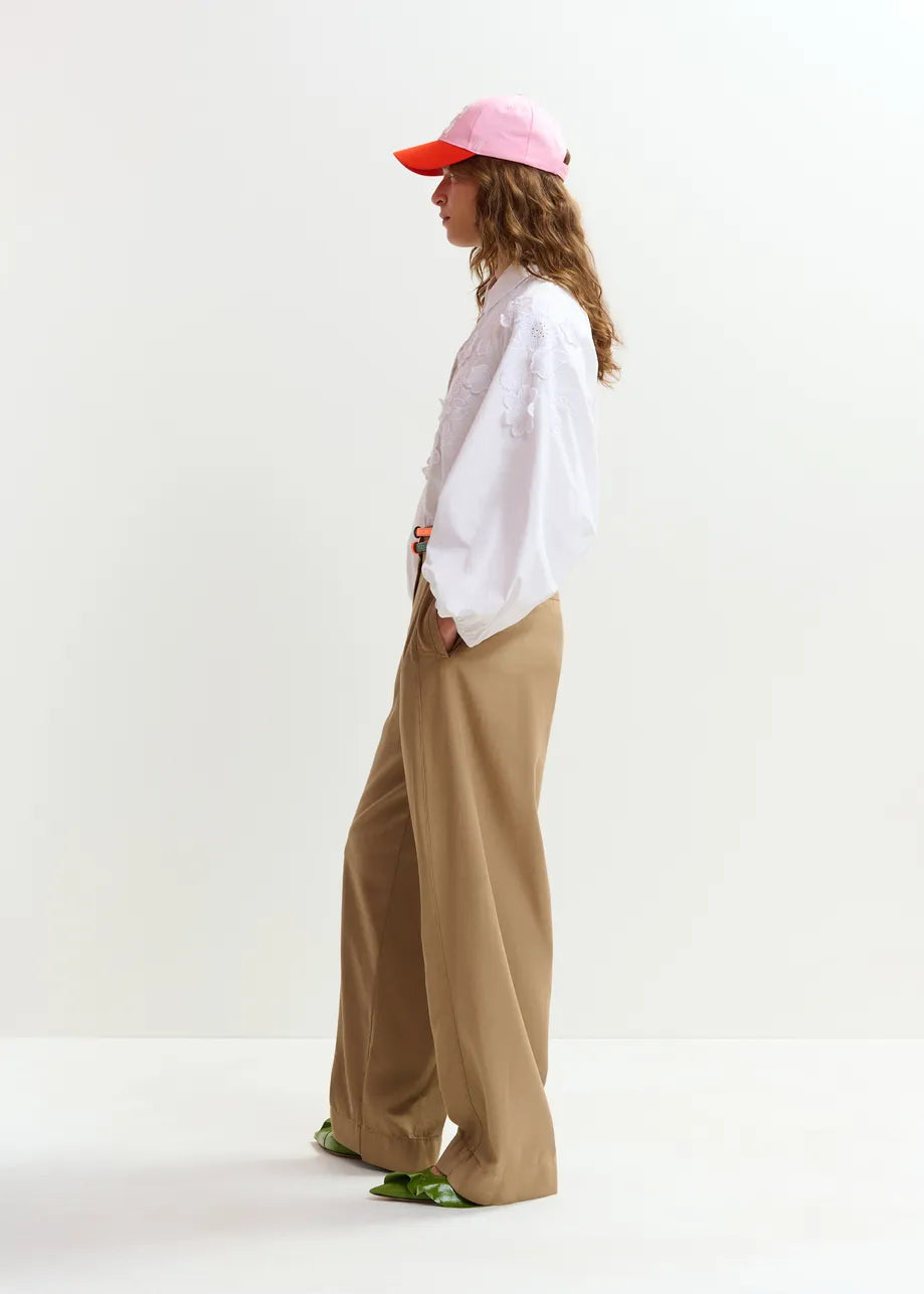 A person stands in profile against a plain background, wearing the Essentiel Antwerp Joella Cotton Flower Shirt in white with balloon sleeves, paired with wide-leg tan pants, a pink and orange cap, and green shoes.