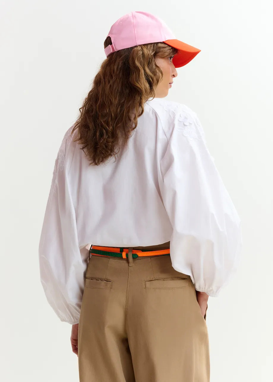 A person wearing a pink and orange cap, khaki pants, a striped belt, and the Joella Cotton Flower Shirt in white by Essentiel Antwerp, facing away from the camera.