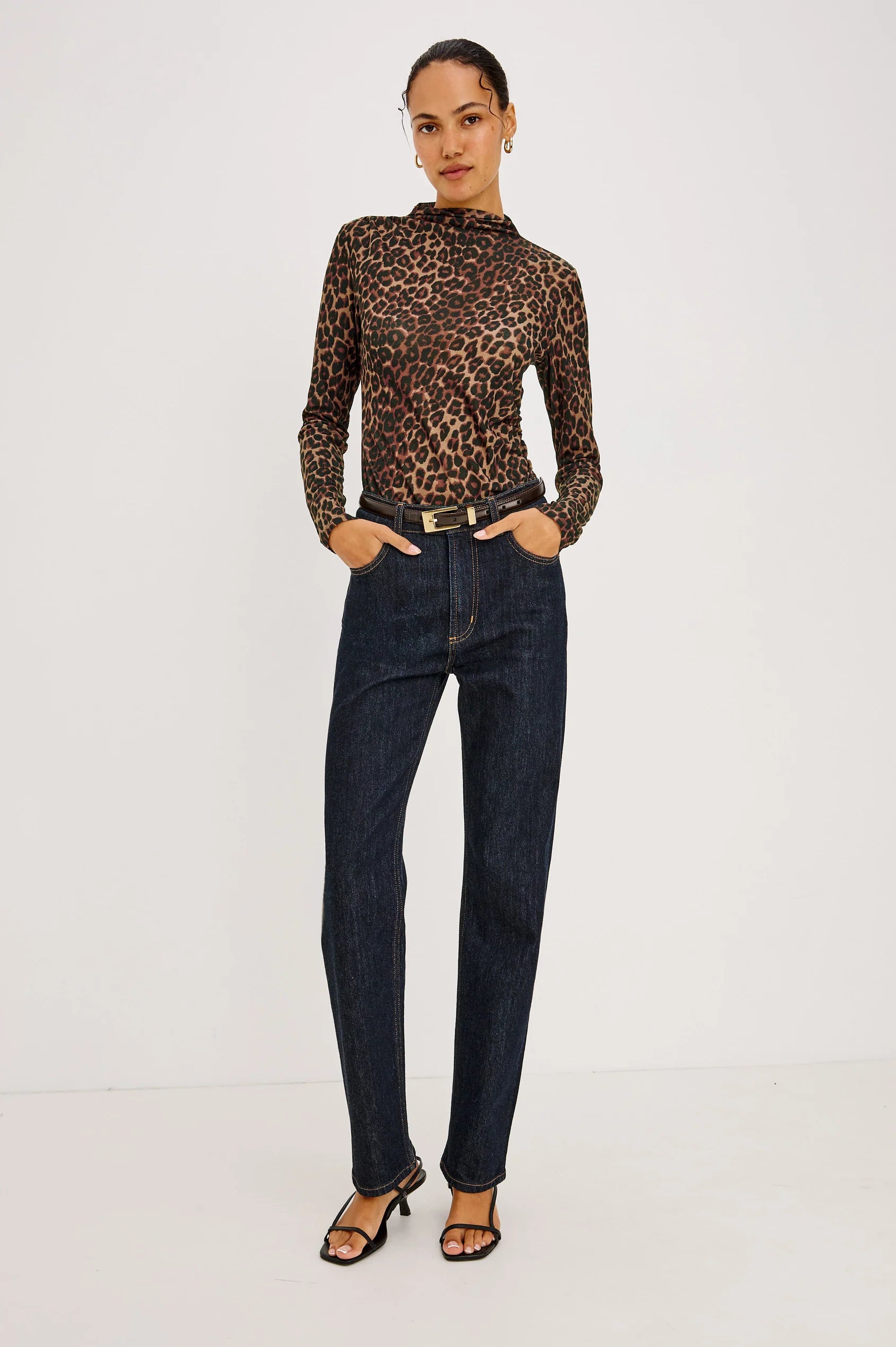 A person wearing a long-sleeved Joelle Top - Brown Nala in a leopard print mesh with a funnel neckline, tucked into dark wash jeans with a black belt and strappy heeled sandals.