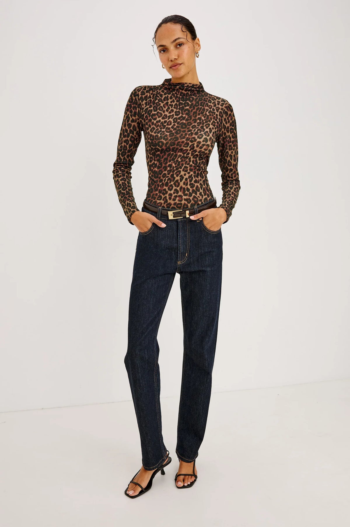 A person wears a brown leopard print Joelle Top - Brown Nala with a funnel neck, tucked into dark wash jeans with a belt and black sandals.