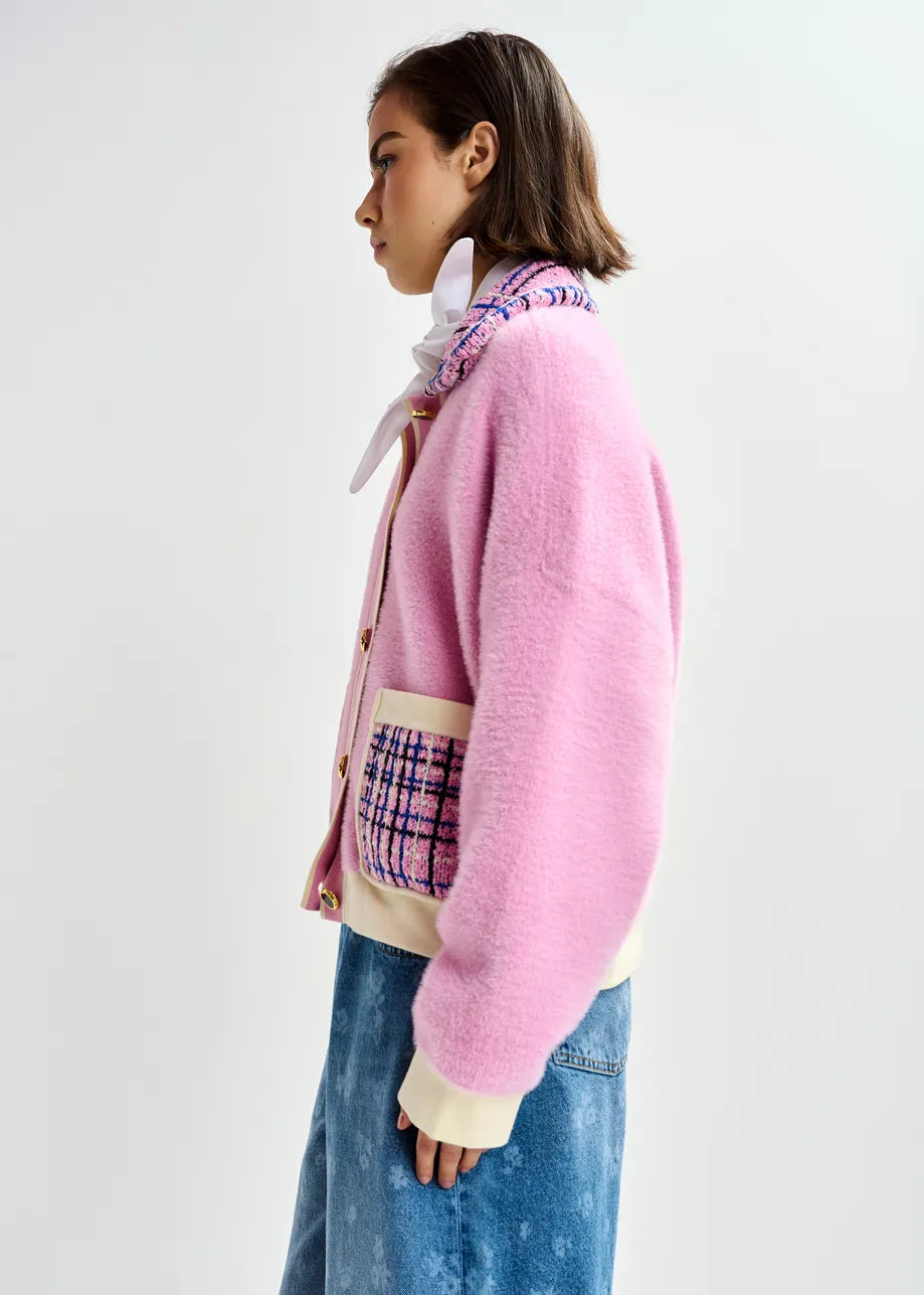 A person stands in profile against a plain background wearing the Essentiel Antwerp Joilu Contrast Knit Cardigan in pink, featuring patterned pockets and collar, paired with a white shirt and blue denim skirt.