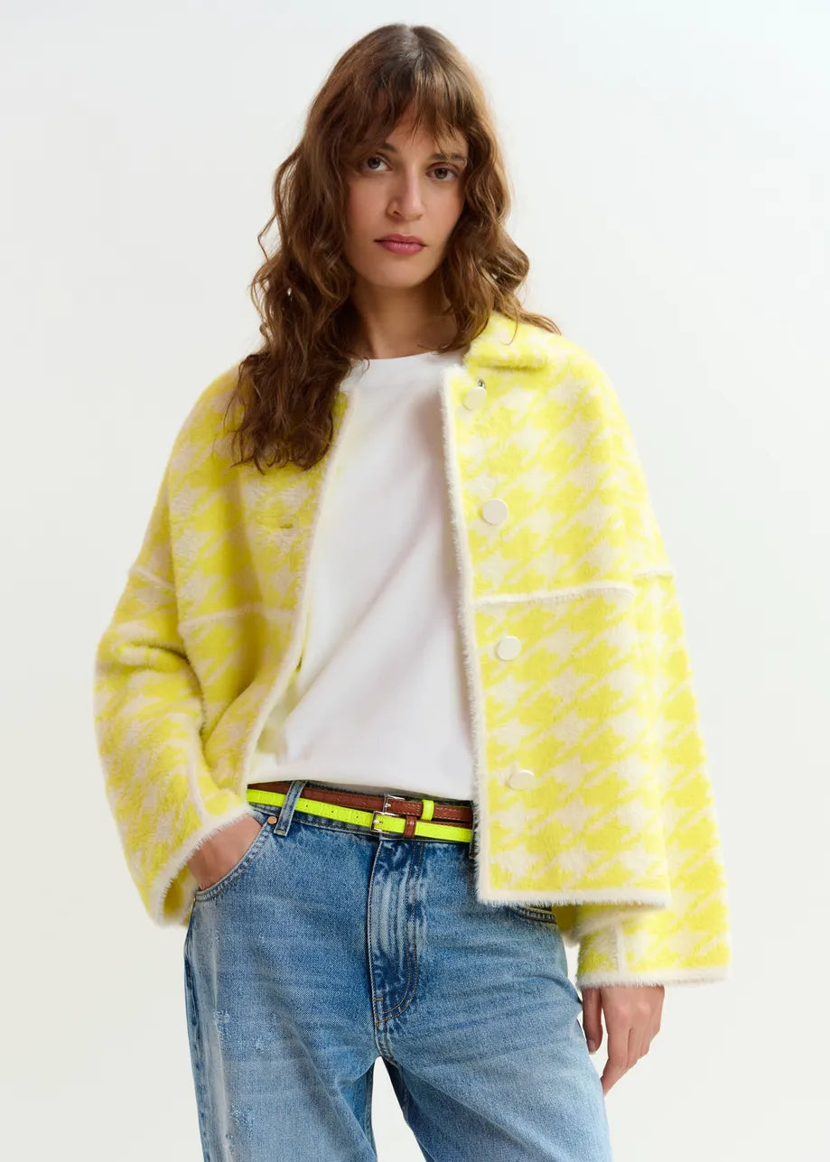 A woman stands against a plain background wearing a white t-shirt, blue jeans, and the Essentiel Antwerp Joking Houndstooth Knit Cardigan in yellow.