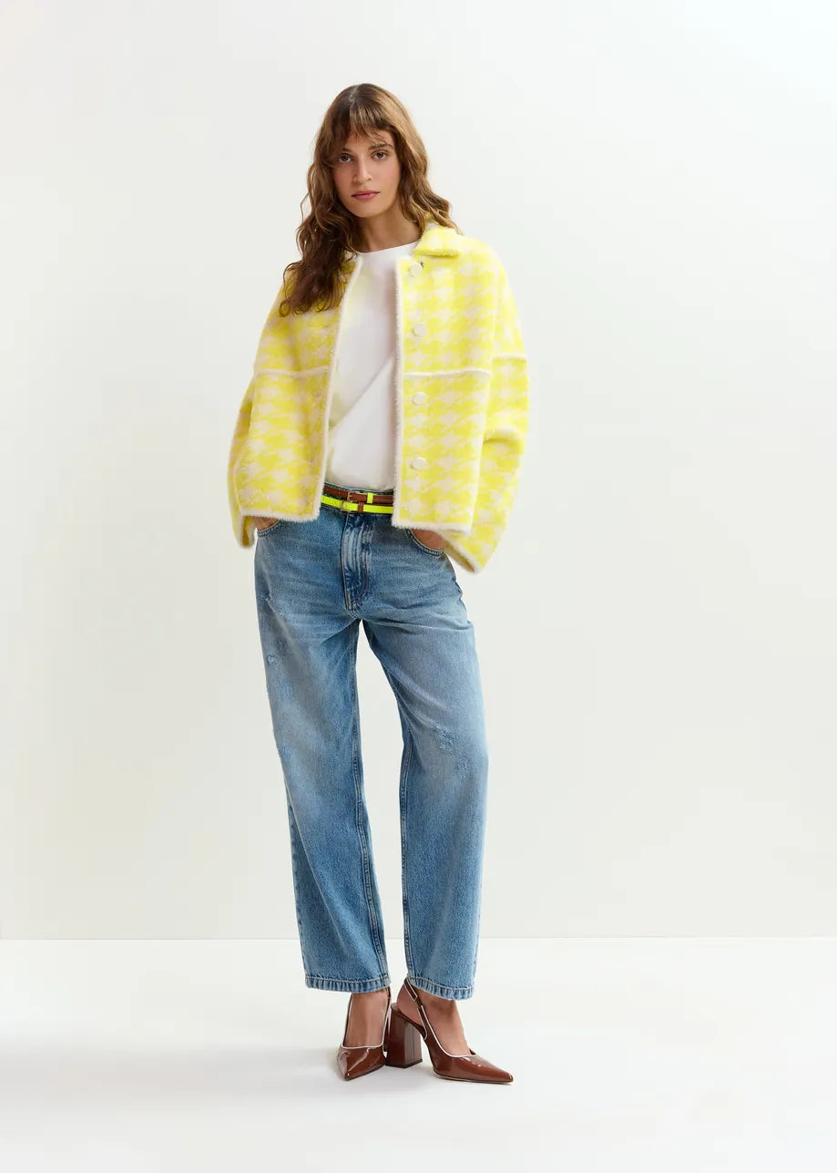 A woman stands against a plain background wearing the Essentiel Antwerp Joking Houndstooth Knit Cardigan in yellow, paired with a white top, blue jeans, and brown heels.