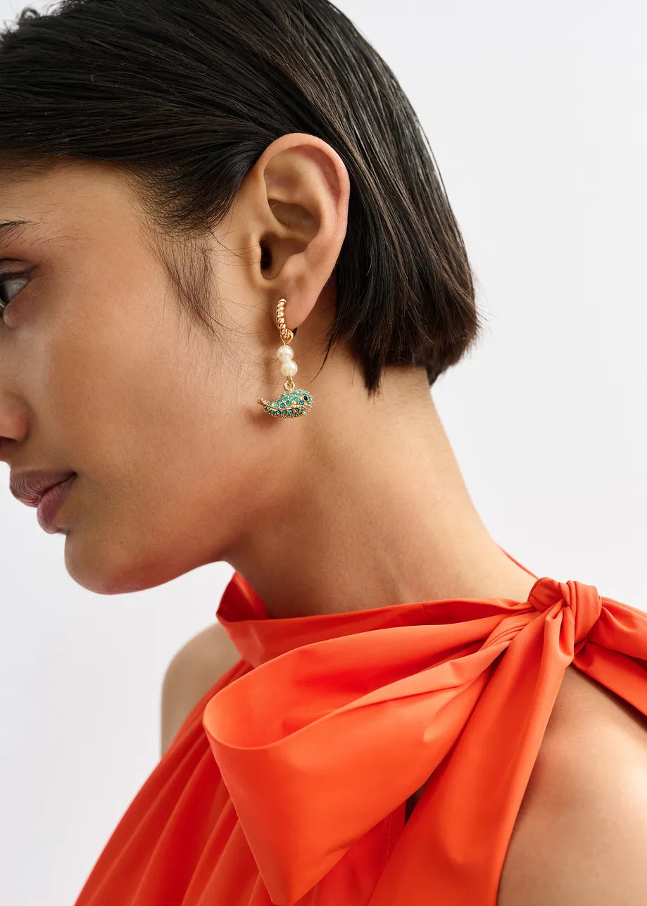 A woman with short dark hair wears a bright orange top and striking mismatched Essentiel Antwerp earrings—one gold drop earring with pearls and turquoise stones, the other the Gold Drop Earrings - Blue Whale.