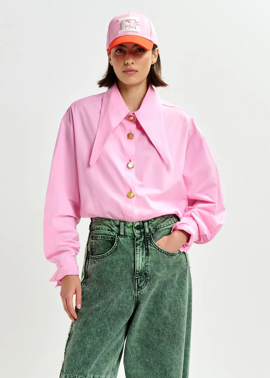 A person wears the Essentiel Antwerp Jomato Pointed Collar Shirt in pink, paired with green jeans and a pink baseball cap, standing against a plain light background.