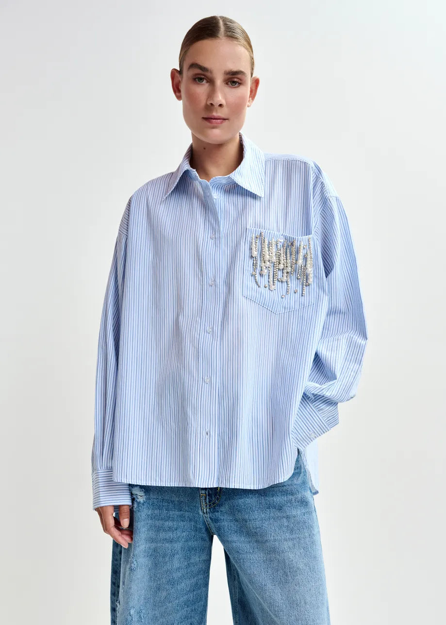 A person wears the Essentiel Antwerp Jommeke Embellished Shirt in blue stripe with blue jeans, standing against a plain background.