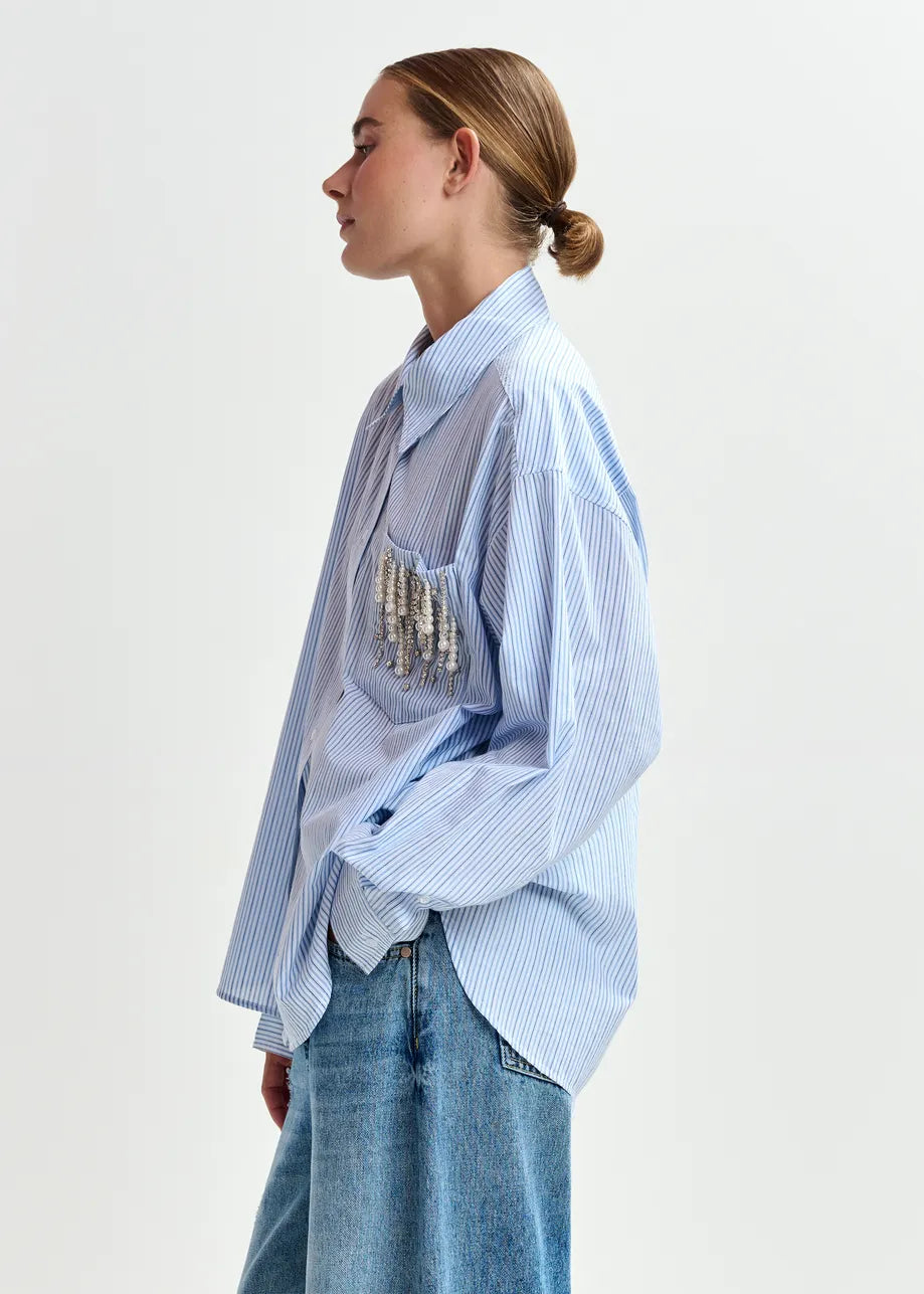 A woman in profile wears the Essentiel Antwerp Jommeke Embellished Shirt - Blue Stripe with blue jeans, standing against a plain background.