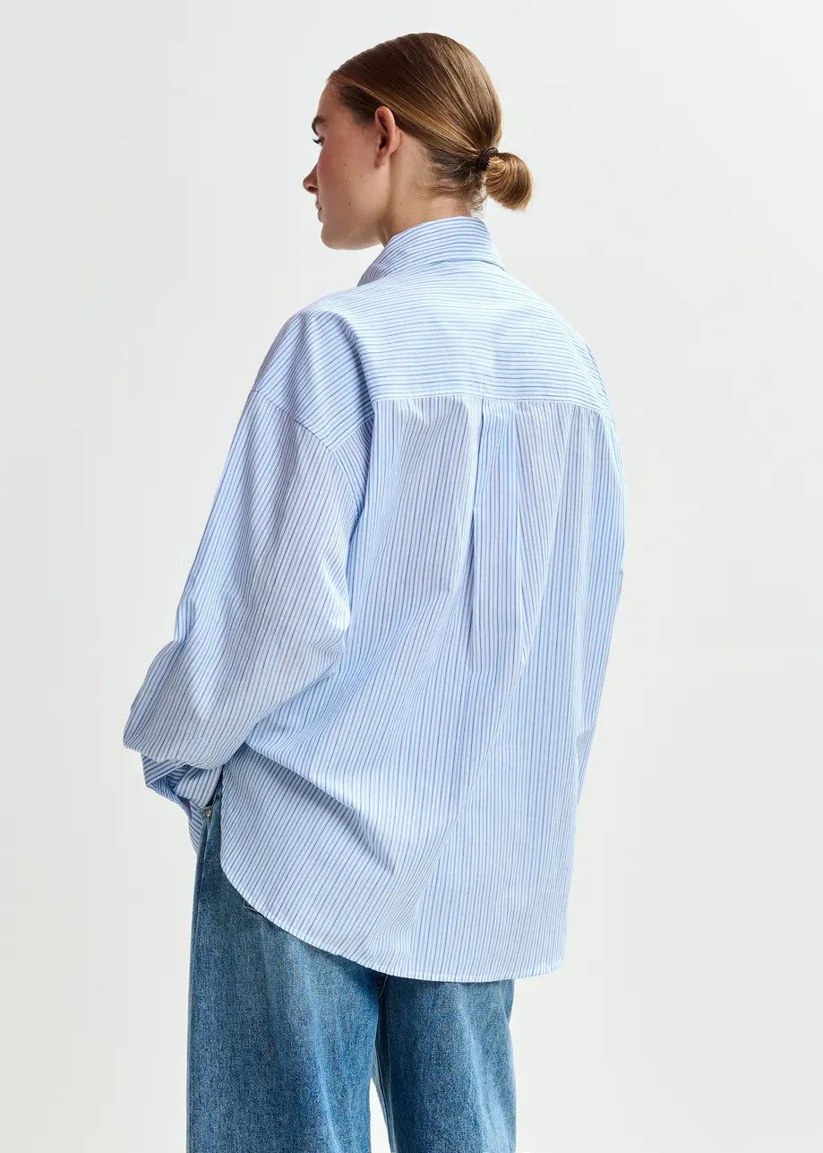Person with hair in a low bun wearing the Essentiel Antwerp Jommeke Embellished Shirt in blue stripes and blue jeans, seen from the back.