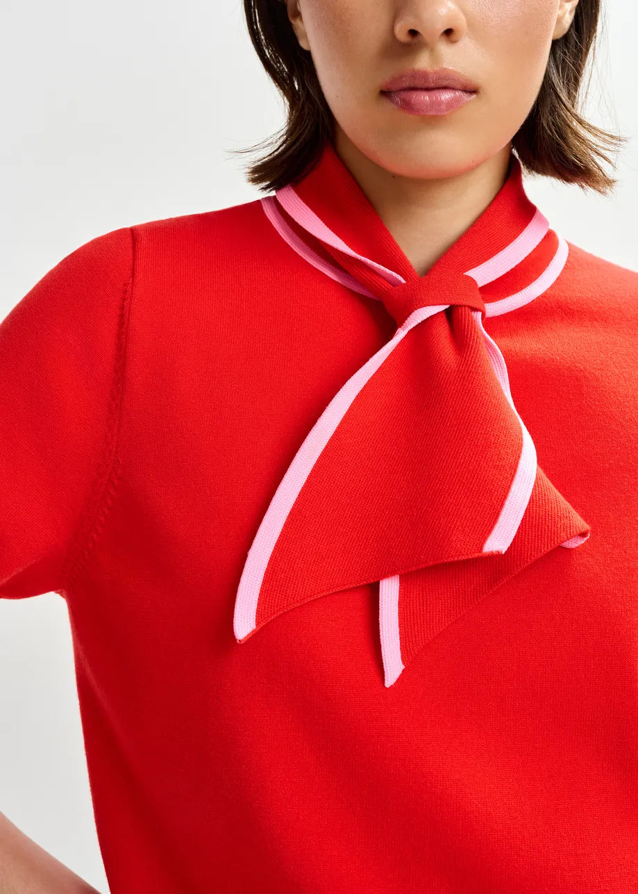 A woman models the Essentiel Antwerp Jonda Knit Polo in red/light pink, featuring a pink-trimmed necktie detail and relaxed fit, shown from the shoulders up against a plain background.