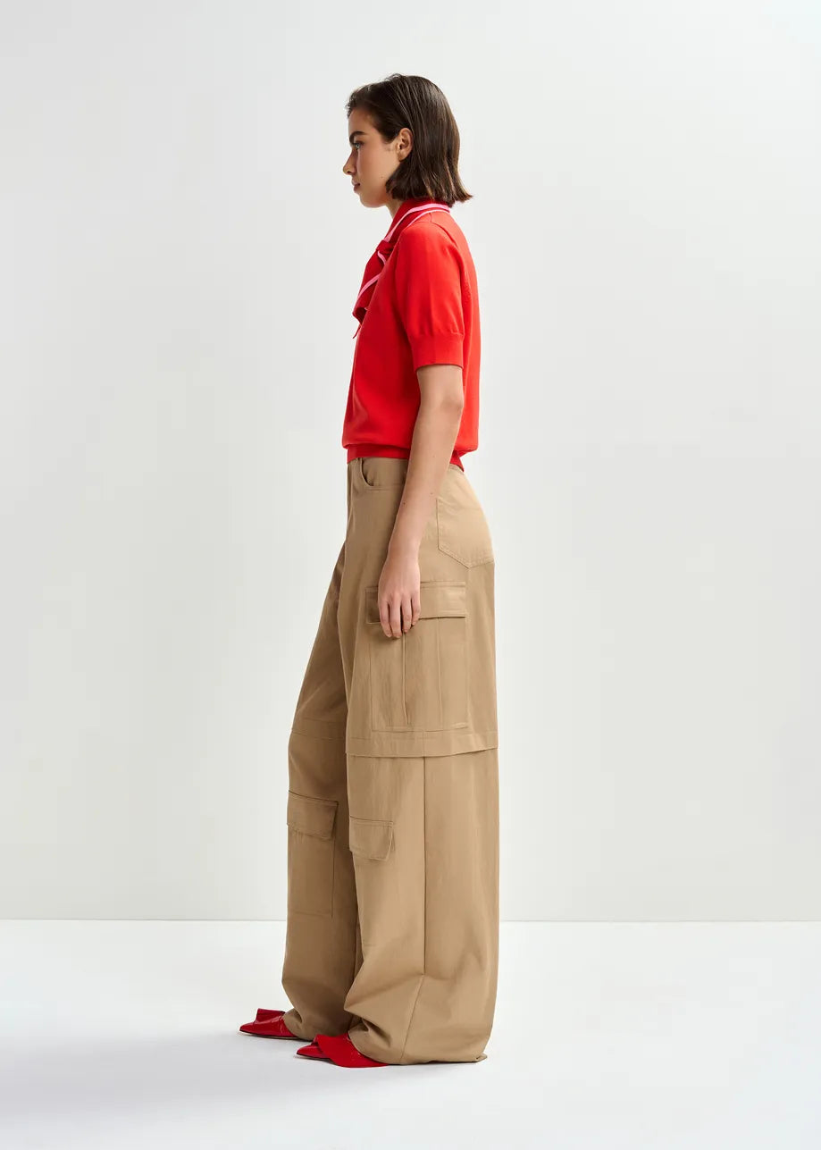 A woman stands in profile against a plain background, wearing Essentiel Antwerp's Jonda Knit Polo in red/light pink, paired with wide-leg tan cargo pants made from recycled polyester and red pointed shoes.