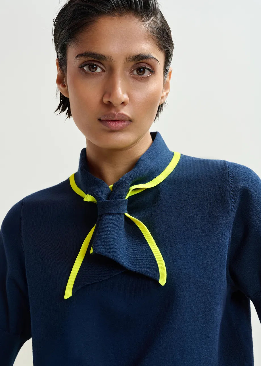A person with short dark hair wears the Essentiel Antwerp Jonda Knit Polo in navy/neon yellow, featuring a relaxed fit, distinctive collar, and neon yellow trim, while looking directly at the camera against a plain background.