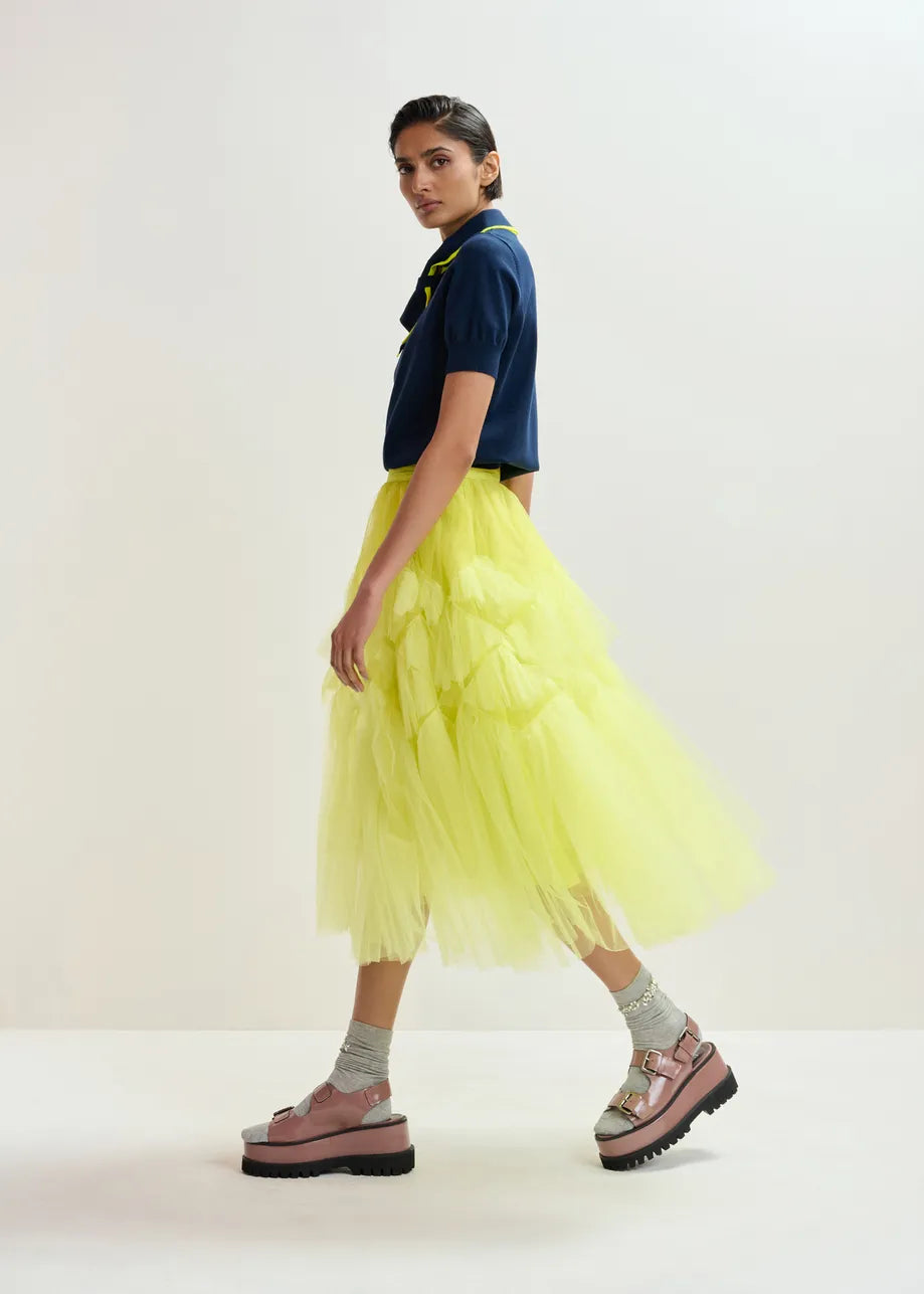 A woman pairs the Essentiel Antwerp Jonda Knit Polo in navy/neon yellow with a yellow tutu skirt, creating a chic and relaxed fit look.