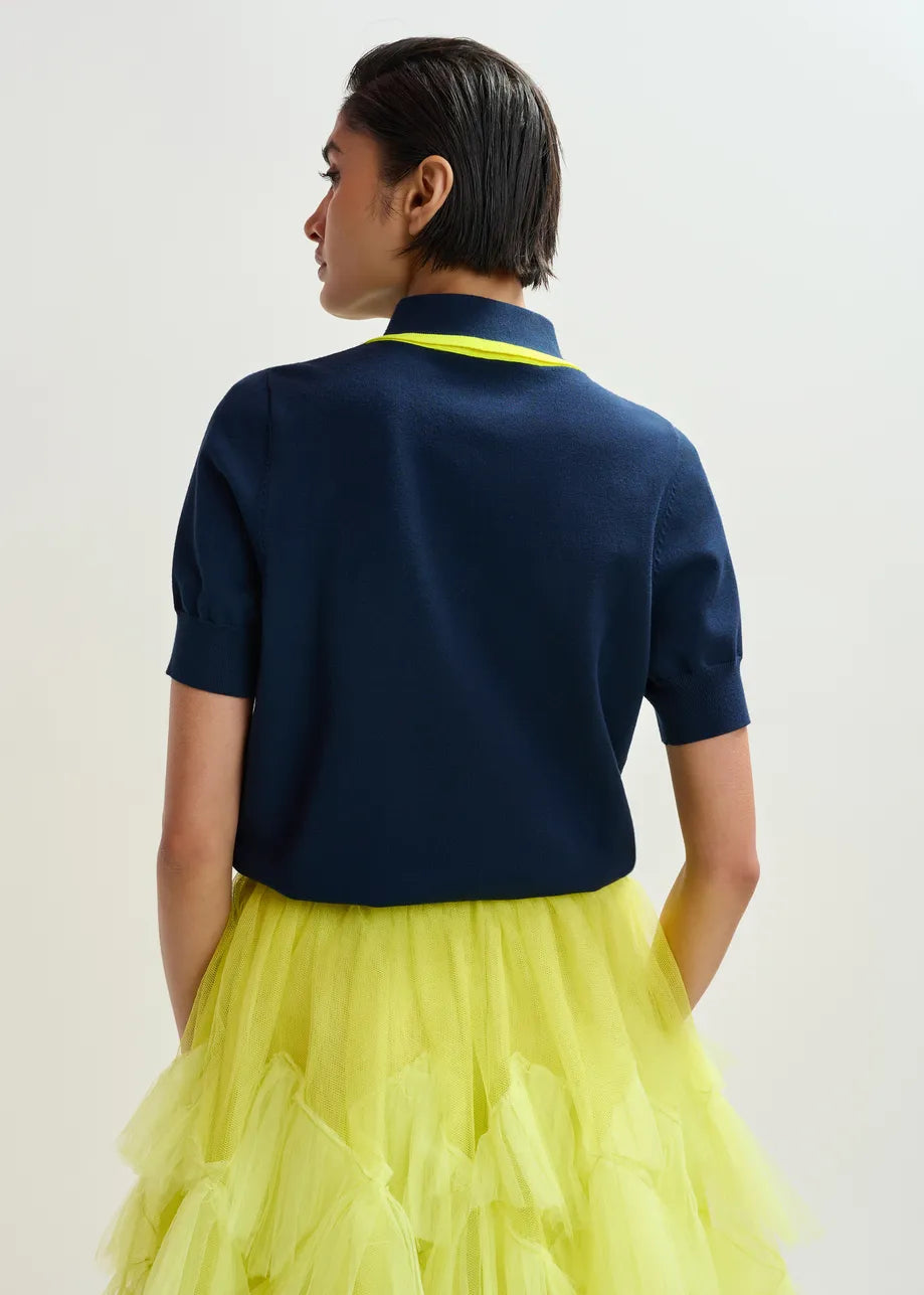 A person faces away from the camera wearing the Essentiel Antwerp Jonda Knit Polo in navy/neon yellow, styled in a relaxed fit with a layered yellow tulle skirt, set against a plain light background.