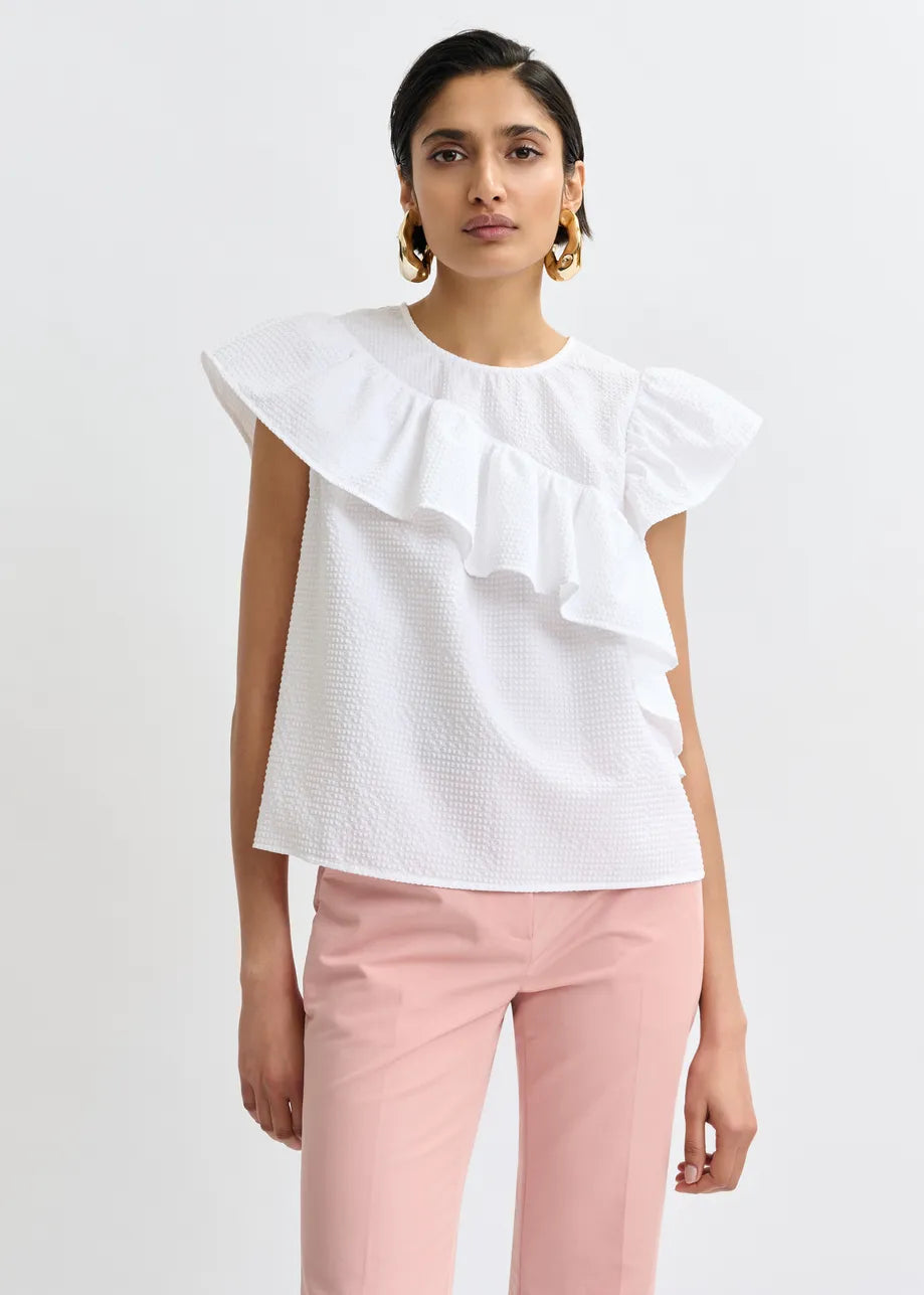 A woman wears the Essentiel Antwerp Joojoo Asymmetric Ruffle Top in white with light pink pants, standing against a plain white background.