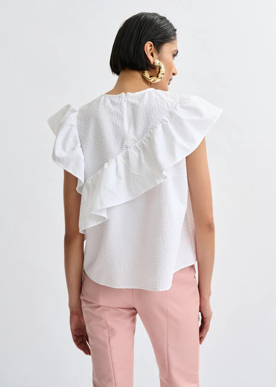 A person faces away, wearing the Essentiel Antwerp Joojoo Asymmetric Ruffle Top in white with crinkle texture and ruffle details, paired with light pink pants, set against a plain light background.