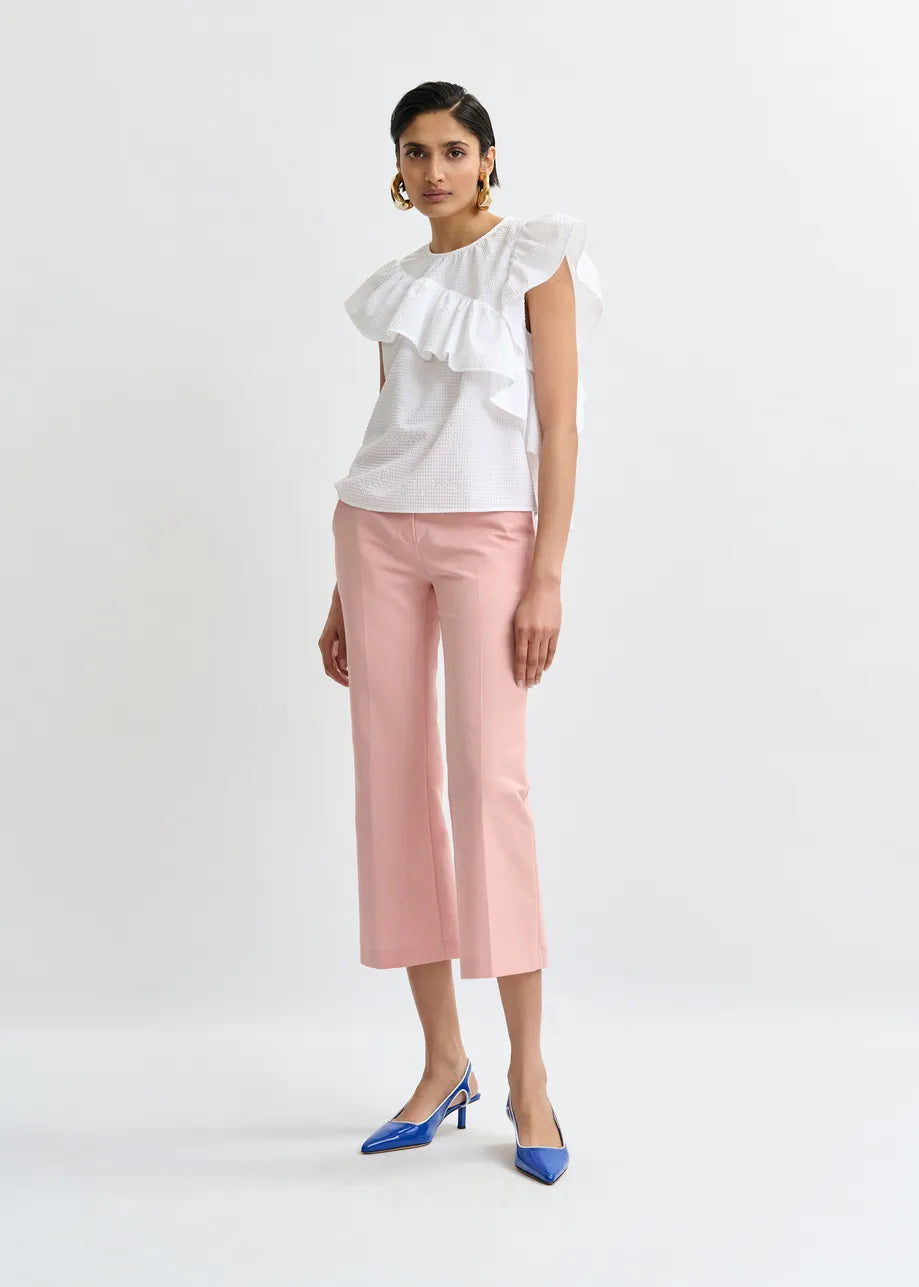 A woman stands against a plain background wearing the Essentiel Antwerp Joojoo Asymmetric Ruffle Top in white, paired with light pink cropped pants and blue pointed-toe heels.