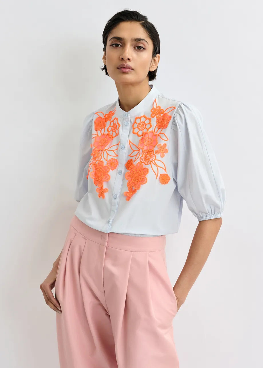 A person wears the Essentiel Antwerp Jorja Flower Shirt in blue with orange floral embroidery, paired with light pink high-waisted pleated trousers, standing against a plain background.