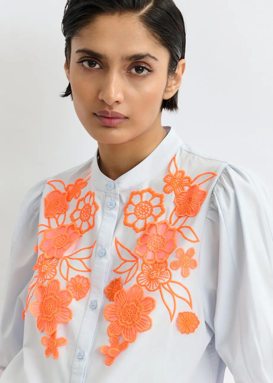 A woman with short dark hair wears the Essentiel Antwerp Jorja Flower Shirt in blue and orange, featuring a mandarin collar and floral embroidery on the front.