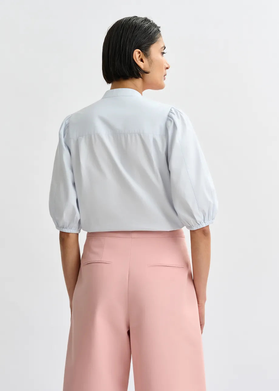 A person with short dark hair is shown from the back, wearing the Essentiel Antwerp Jorja Flower Shirt in blue/orange and high-waisted pink trousers against a plain background.
