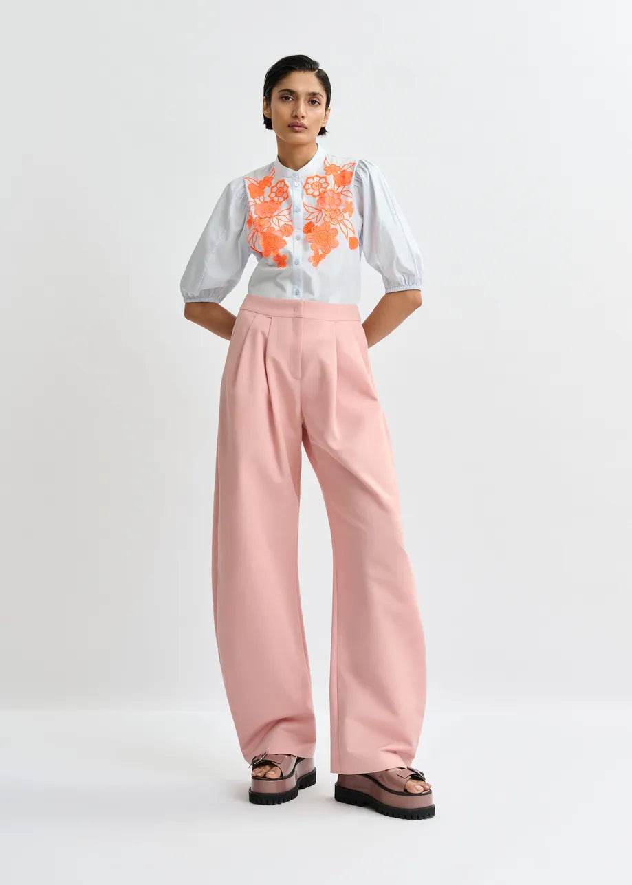 A woman stands against a white background wearing the Essentiel Antwerp Jorja Flower Shirt in blue and orange, featuring floral embroidery and a mandarin collar, paired with high-waisted pink wide-leg pants and platform sandals.