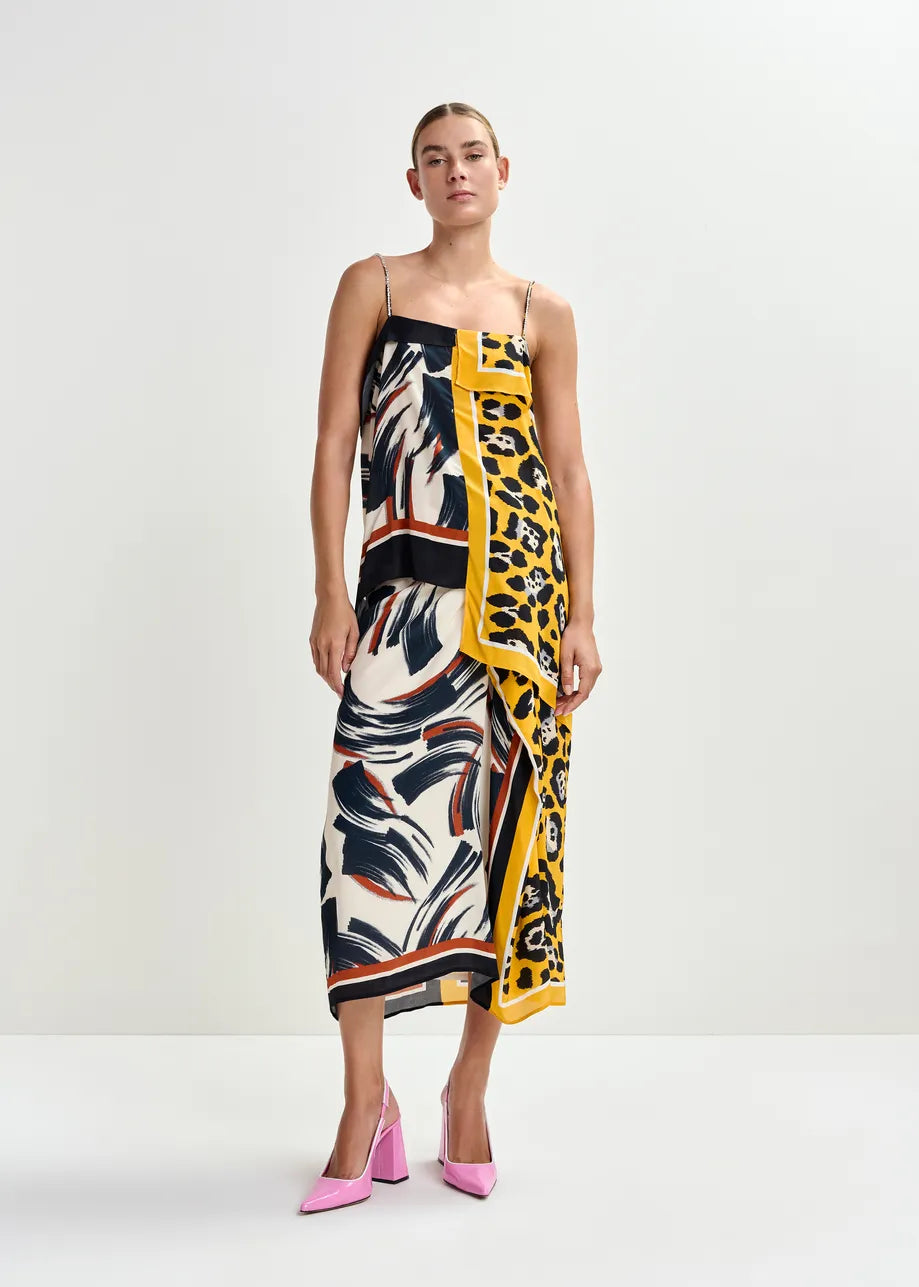 A woman stands in the Essentiel Antwerp Juanita Drape Midi Skirt in ochre, featuring a relaxed fit with abstract, leopard, and tropical prints, paired with pink pointed-toe heels against a plain background.