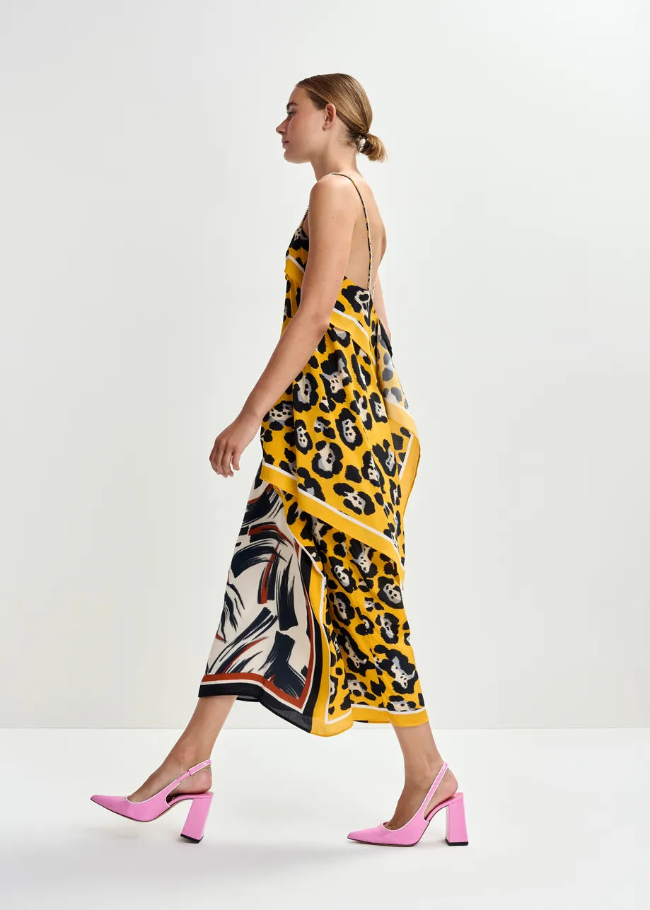 A woman wearing the Essentiel Antwerp Juanita Drape Midi Skirt in ochre walks against a plain white background, styling it with pink block-heeled shoes.