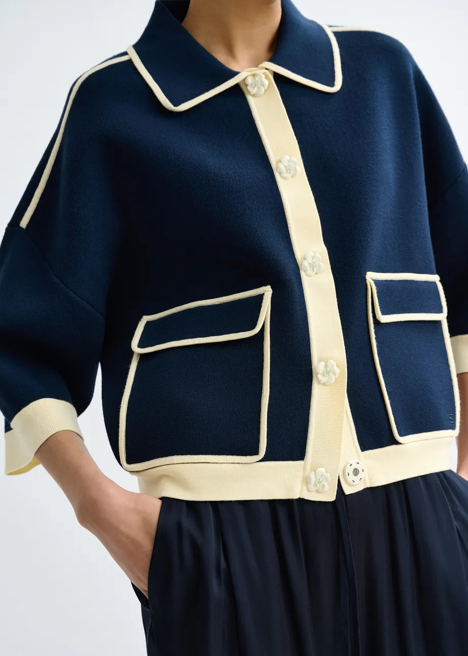 A person wears the Essentiel Antwerp Jugly Contrast Knit Cardigan in navy with cream trim and flower snap buttons, featuring cropped 3/4 sleeves, paired with black pleated pants and hands in pockets.