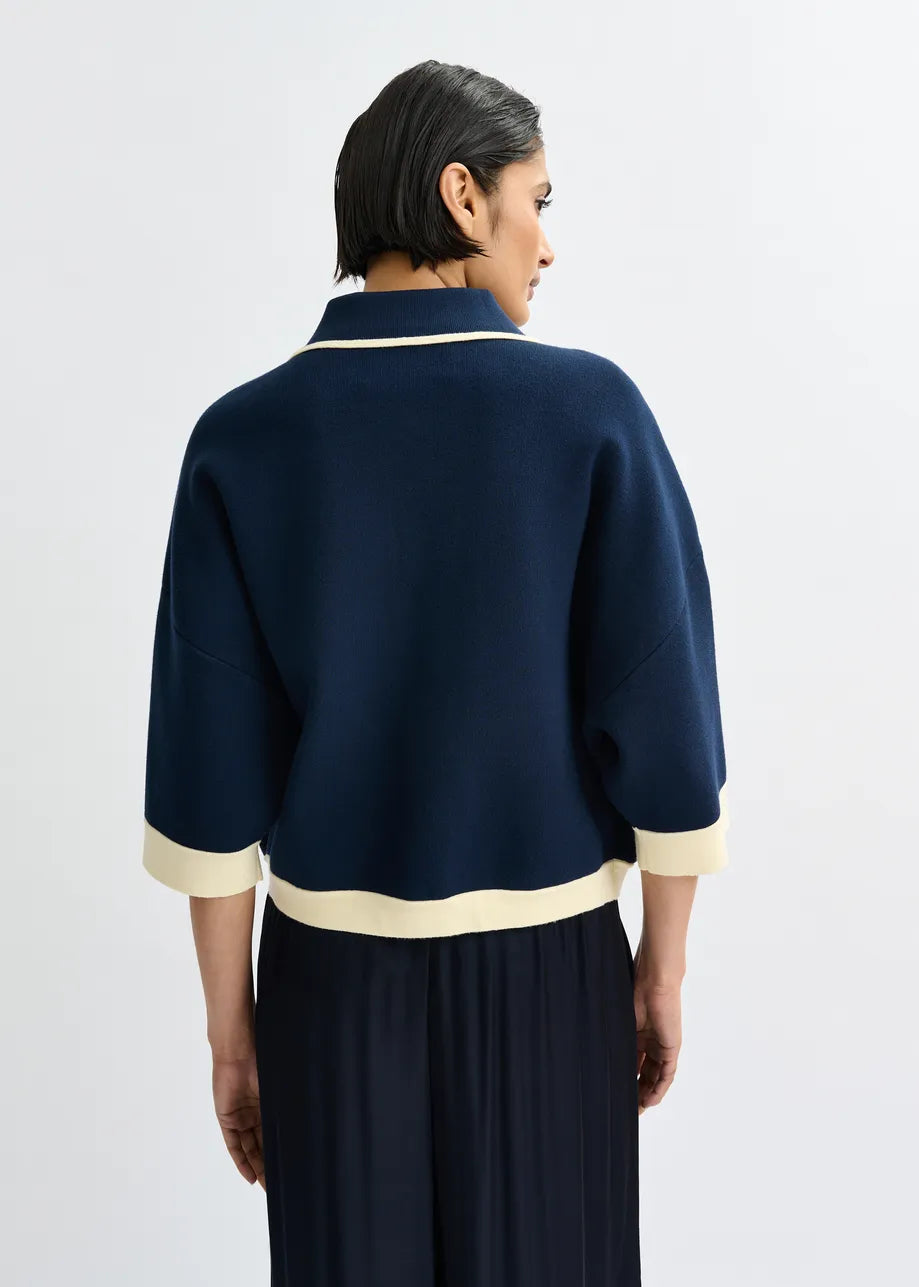 A person with short dark hair is seen from the back wearing the Essentiel Antwerp Jugly Contrast Knit Cardigan in navy with cream trim and cropped 3/4 sleeves, paired with a dark pleated skirt, against a plain light background.