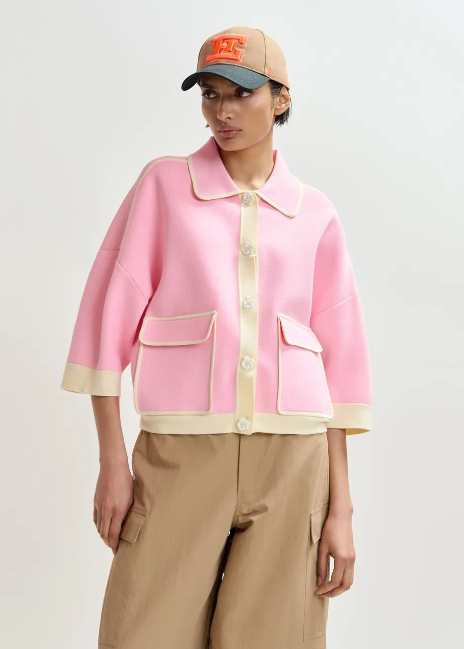 A person wearing the Essentiel Antwerp Jugly Contrast Knit Cardigan in pink with cream trim and flower snap buttons, paired with tan cargo pants and a tan baseball cap featuring an orange "H".