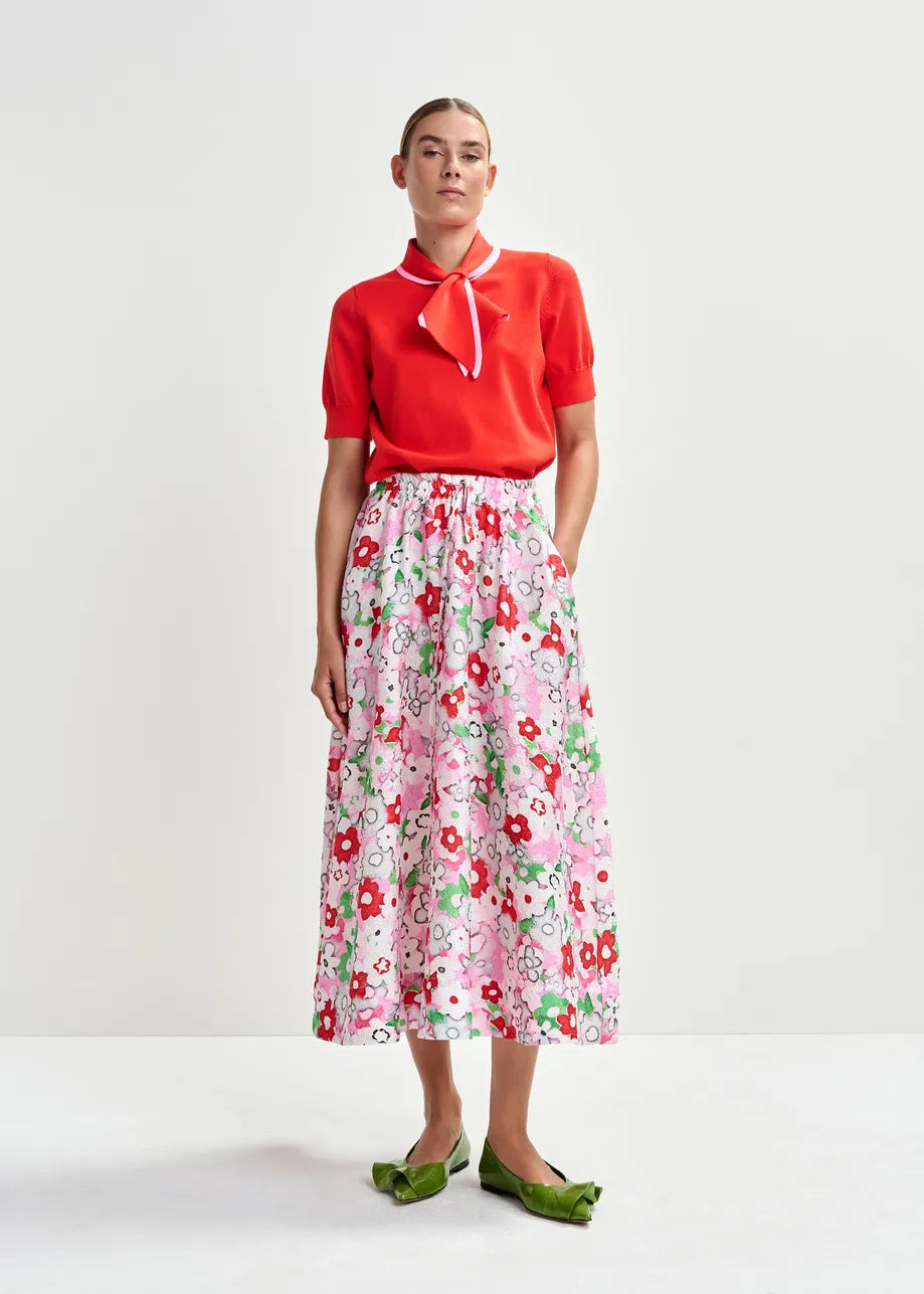 A person stands against a plain background wearing a red short-sleeve top with a white collar tie, the Essentiel Antwerp Julianne Midi Skirt - Floral, and green flat shoes.