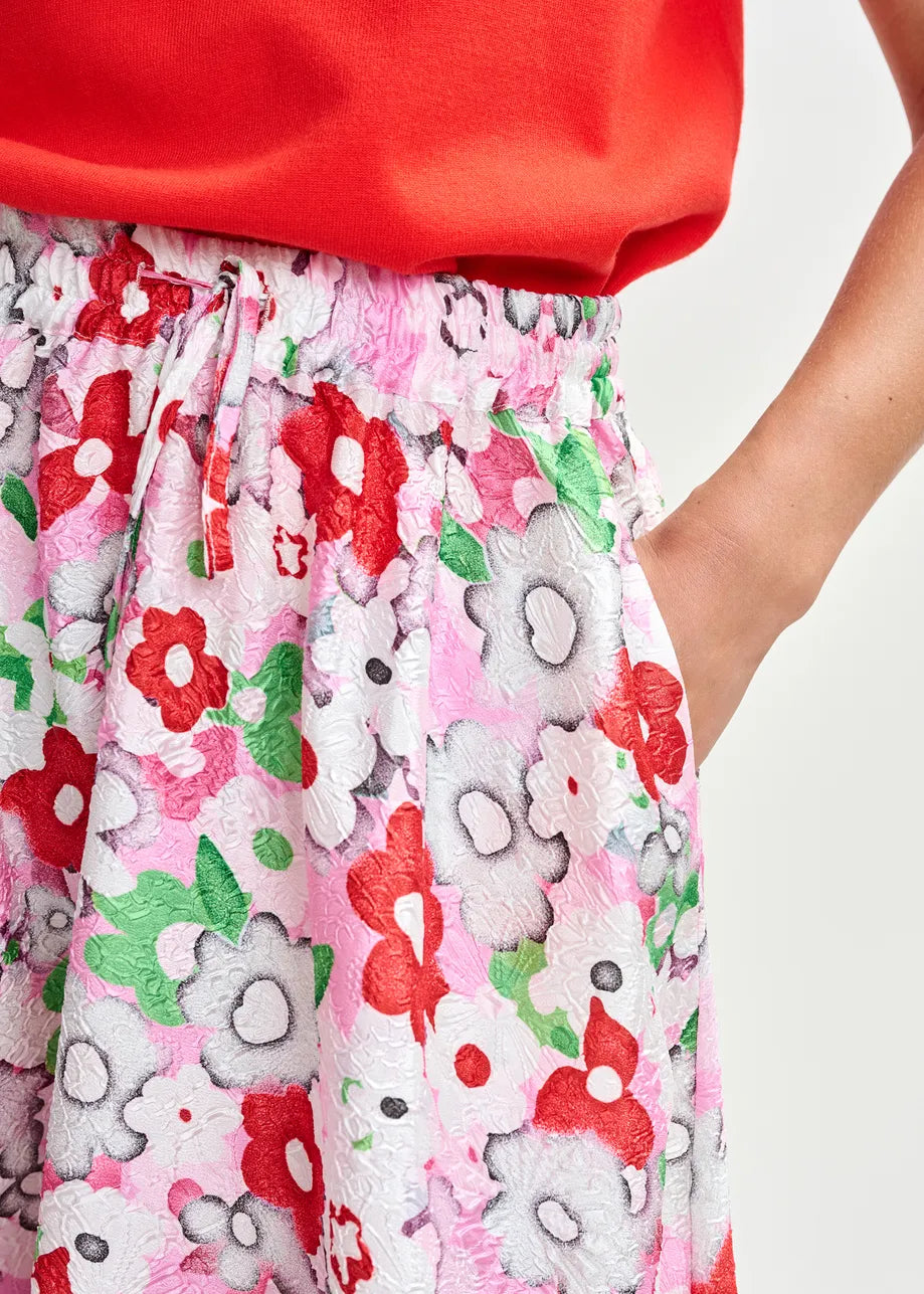 Close-up of a person wearing Essentiel Antwerp’s Julianne Midi Skirt - Floral, featuring a pink, red, white, and green jacquard print, A-line silhouette, drawstring waist, and pockets; paired with a red top.