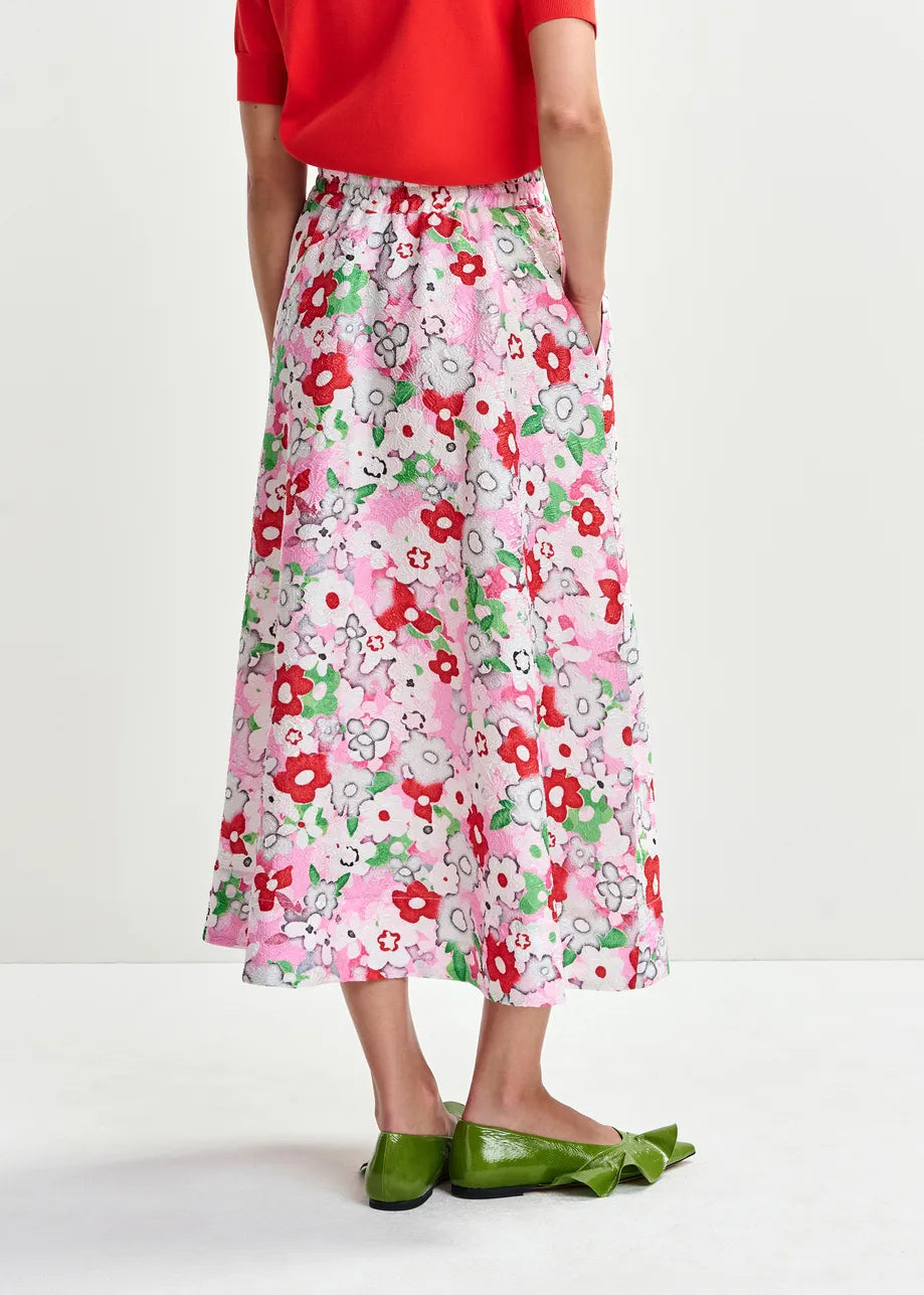 Someone wearing a red top and Essentiel Antwerp's Julianne Midi Skirt - Floral, featuring an A-line silhouette, stands with hands in skirt pockets and green flats, facing away from the camera.