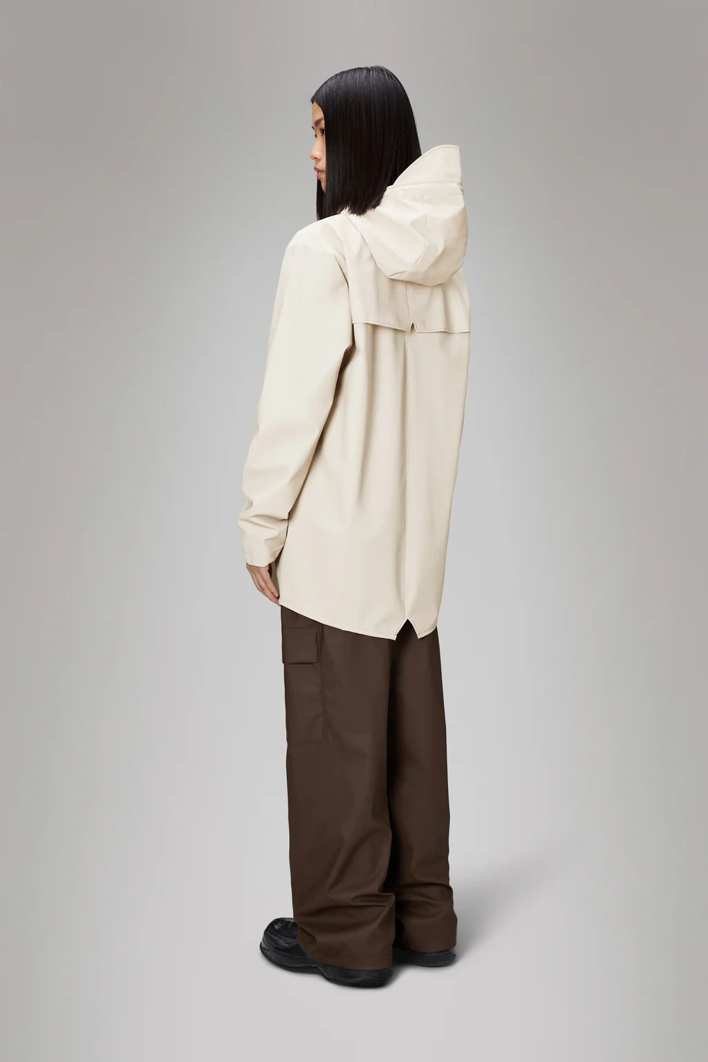 A person with long dark hair, wearing the Rains Jacket W3 in beige and brown wide-leg pants, stands against a plain gray background, facing slightly away from the camera.