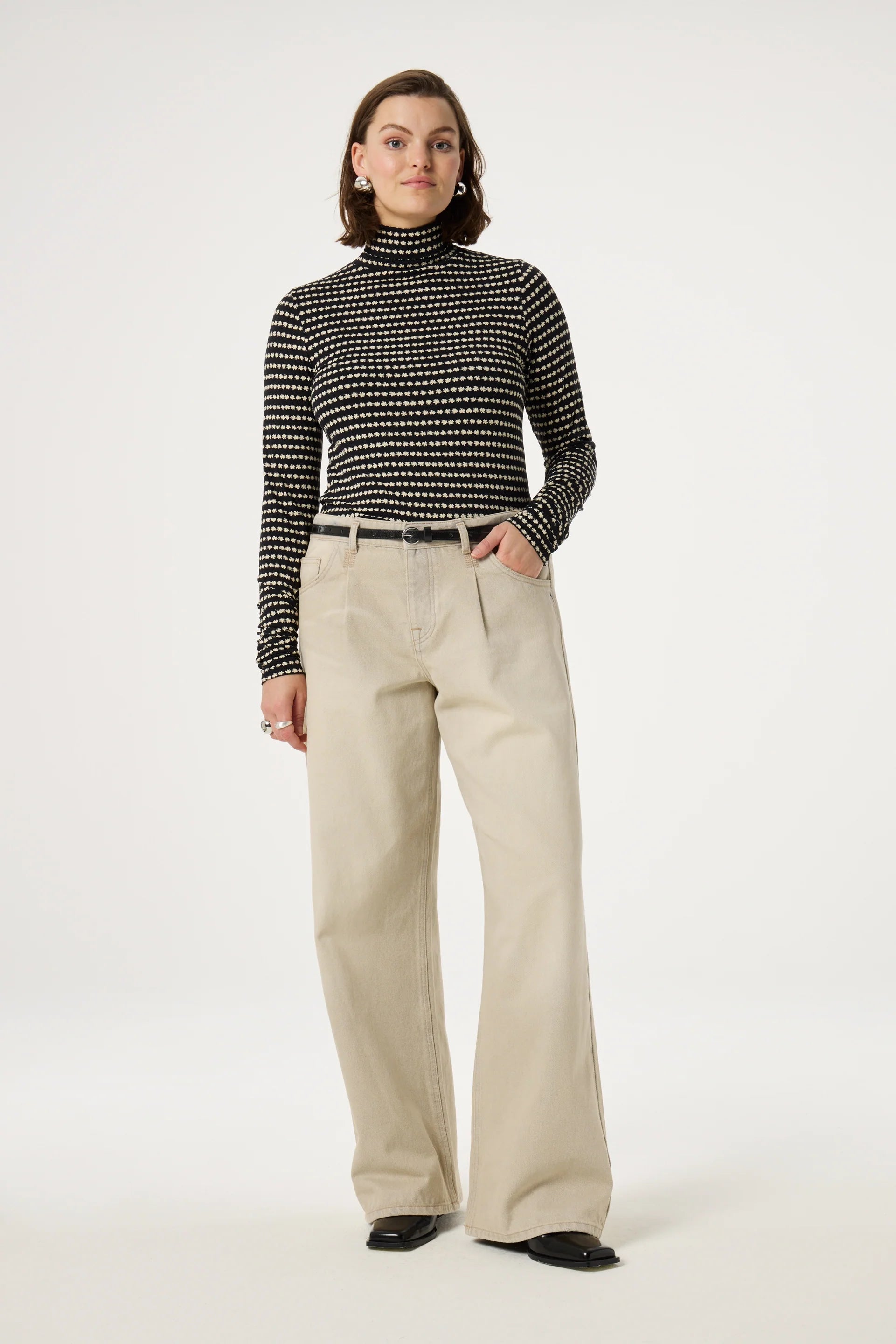 A person wearing the Jade Top - Black by Fabienne Chapot, featuring a chic black and white flower pattern, paired with beige wide-leg pants stands confidently in a neutral pose against a plain background.